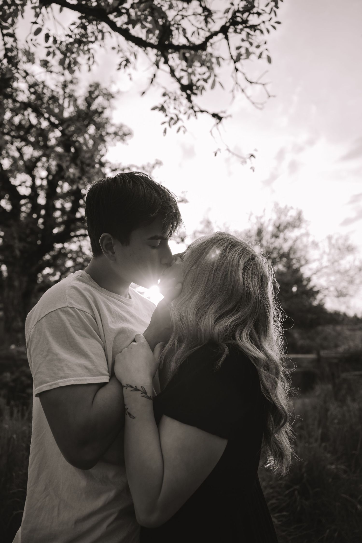 Black and white photo of a couple sharing a romantic kiss outdoors under trees with sunlight glowing behind them.
