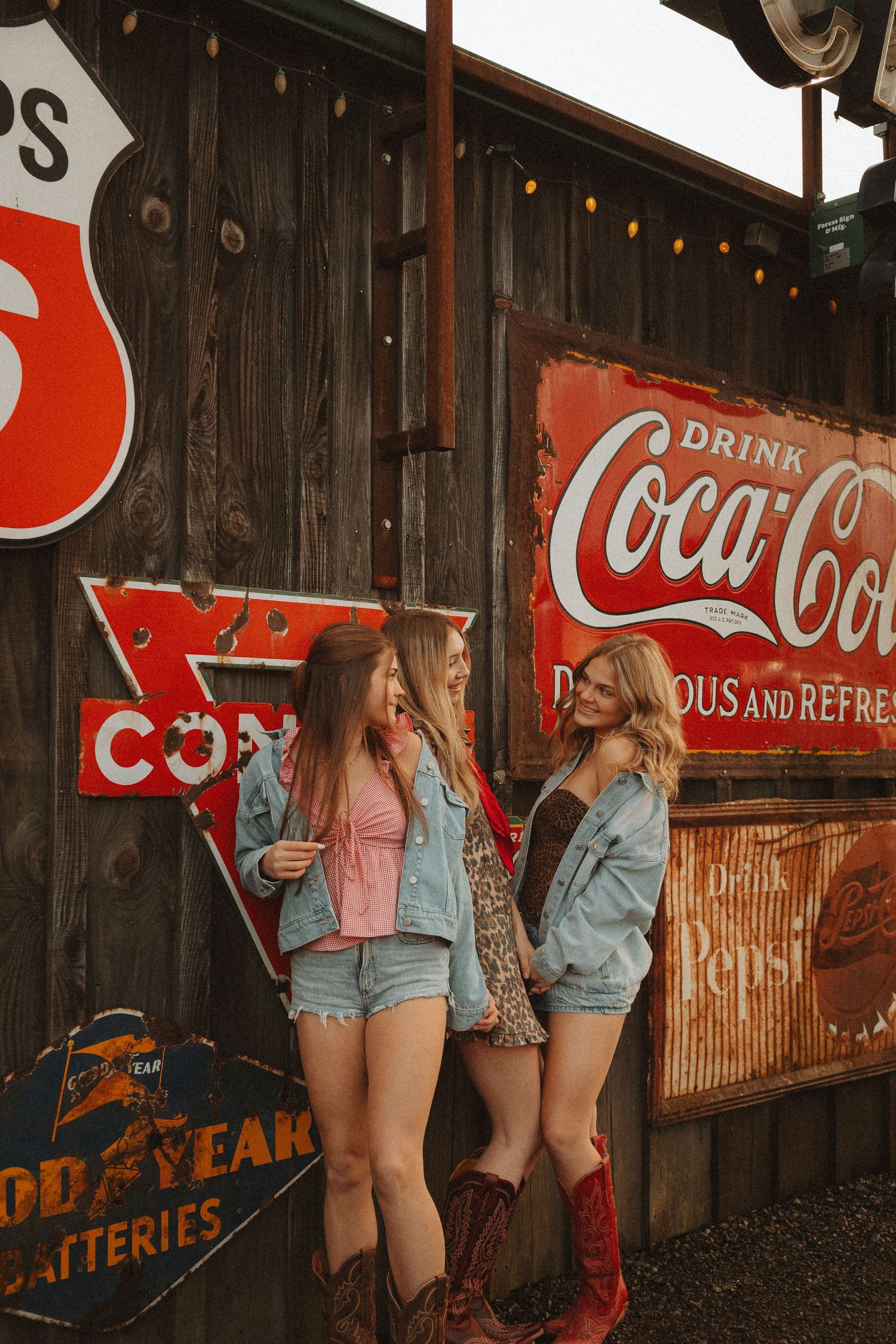 Three women in denim and boots laughing outside rustic barn wall with vintage Coca-Cola and Pepsi signs.