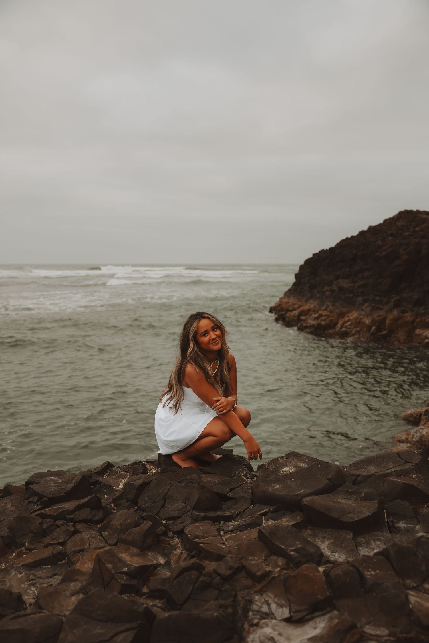 Young woman in white dress sitting on coastal rocks with ocean waves and rocky cliffs in background.