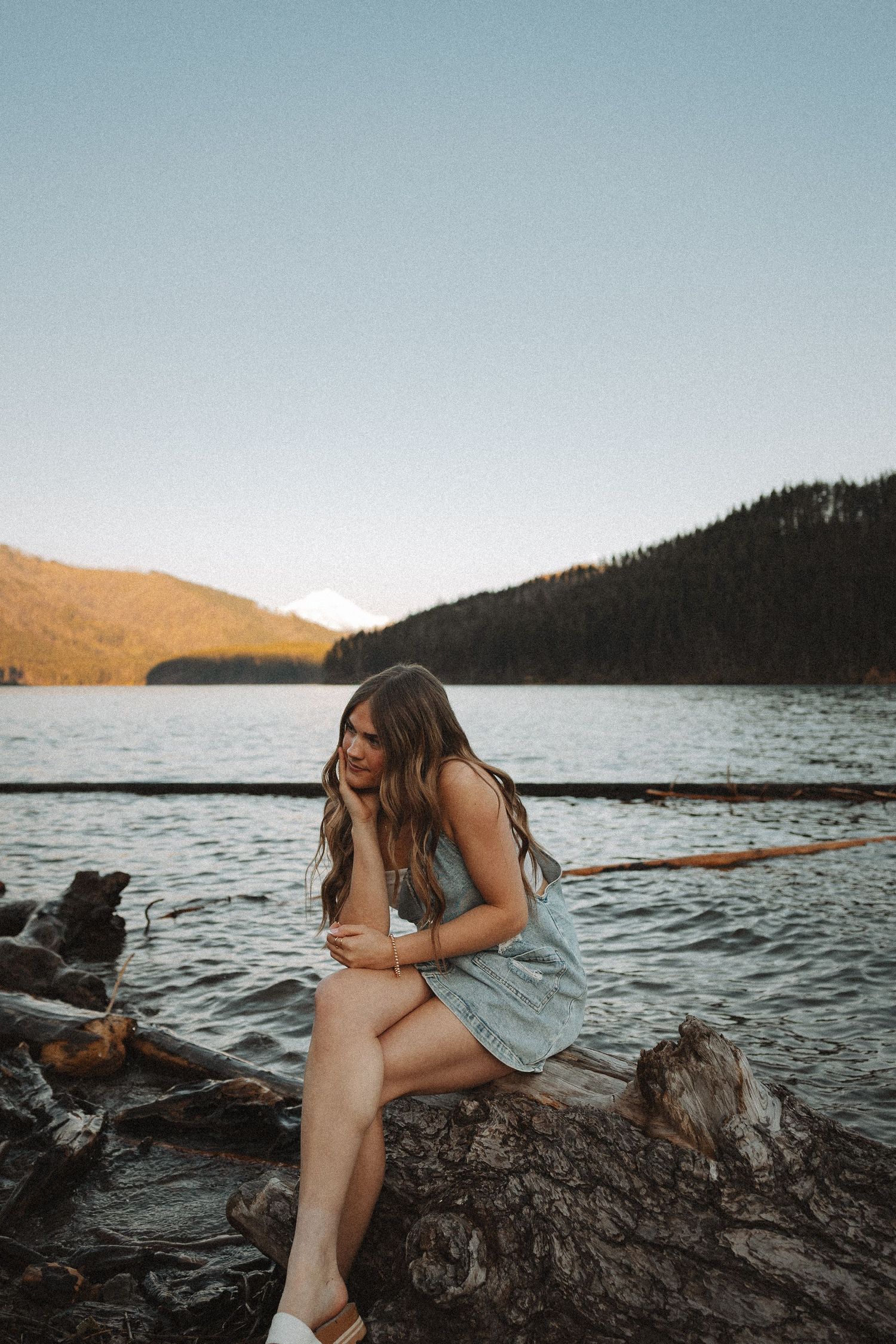 Young woman in light blue dress sitting on driftwood logs by a calm mountain lake at sunset.
