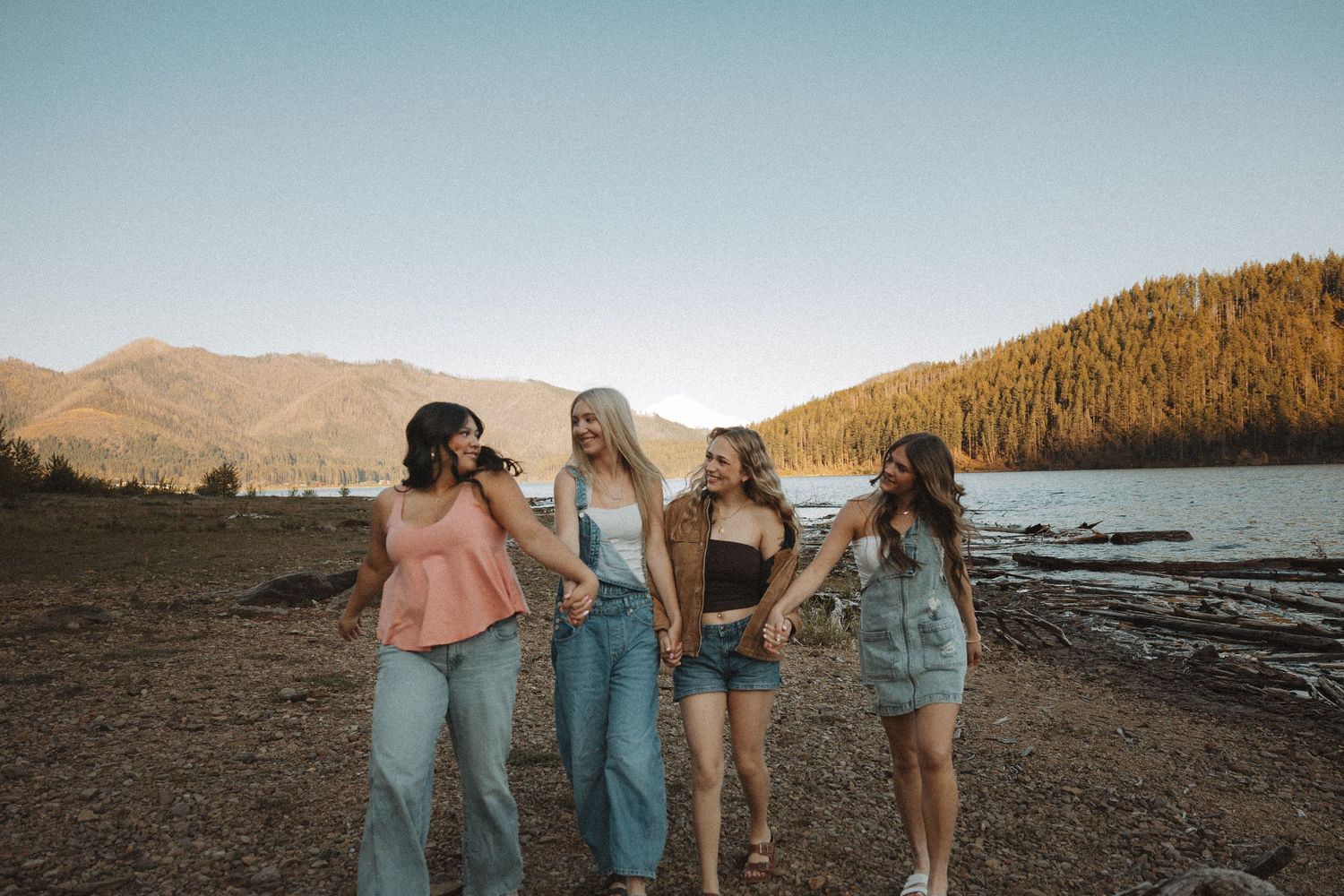 Four young women laughing and walking together along a scenic mountain lake shore at golden hour.