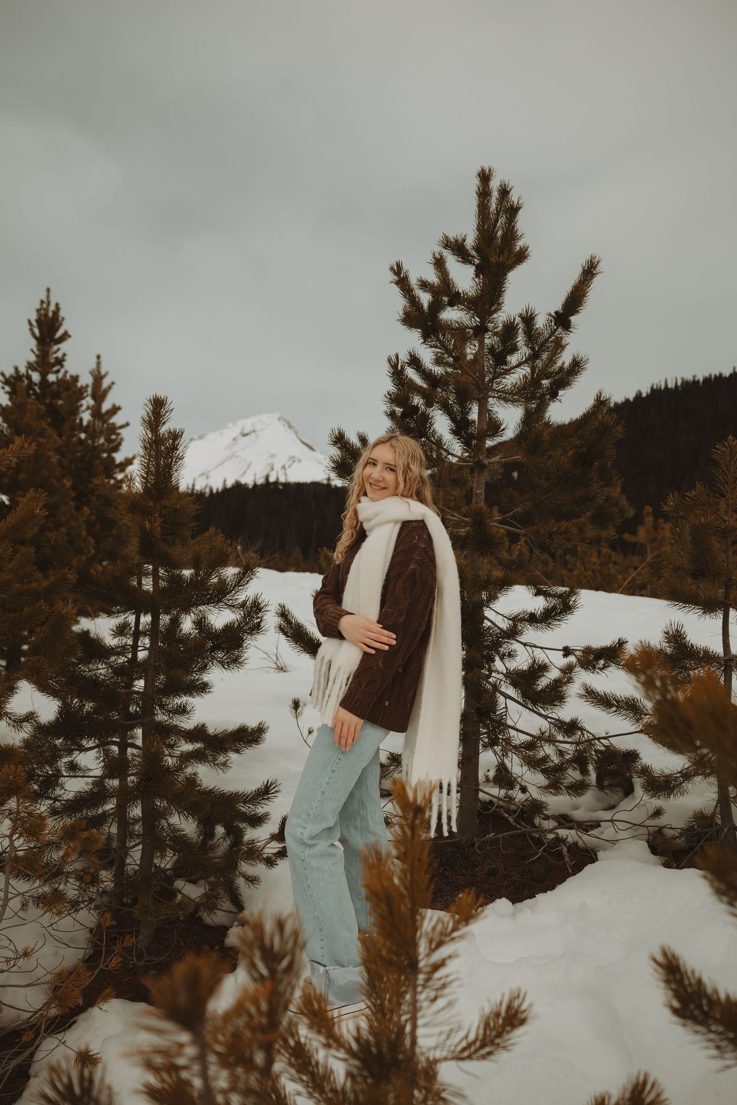 Smiling blonde woman in brown jacket and white scarf stands among snow-dusted pine trees in winter.