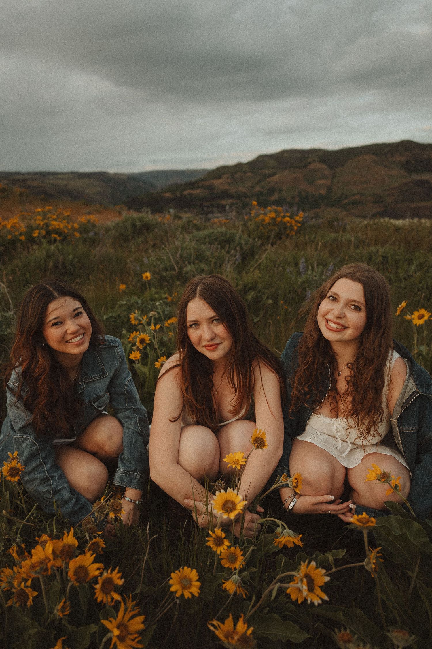Three young women smiling and crouching in a field of yellow wildflowers with rolling hills in the background.