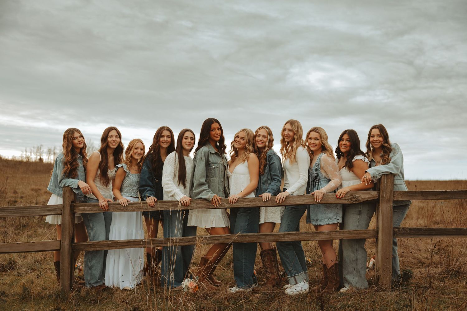 A group in white dresses and brown outfits pose together along a rustic wooden fence at sunset.