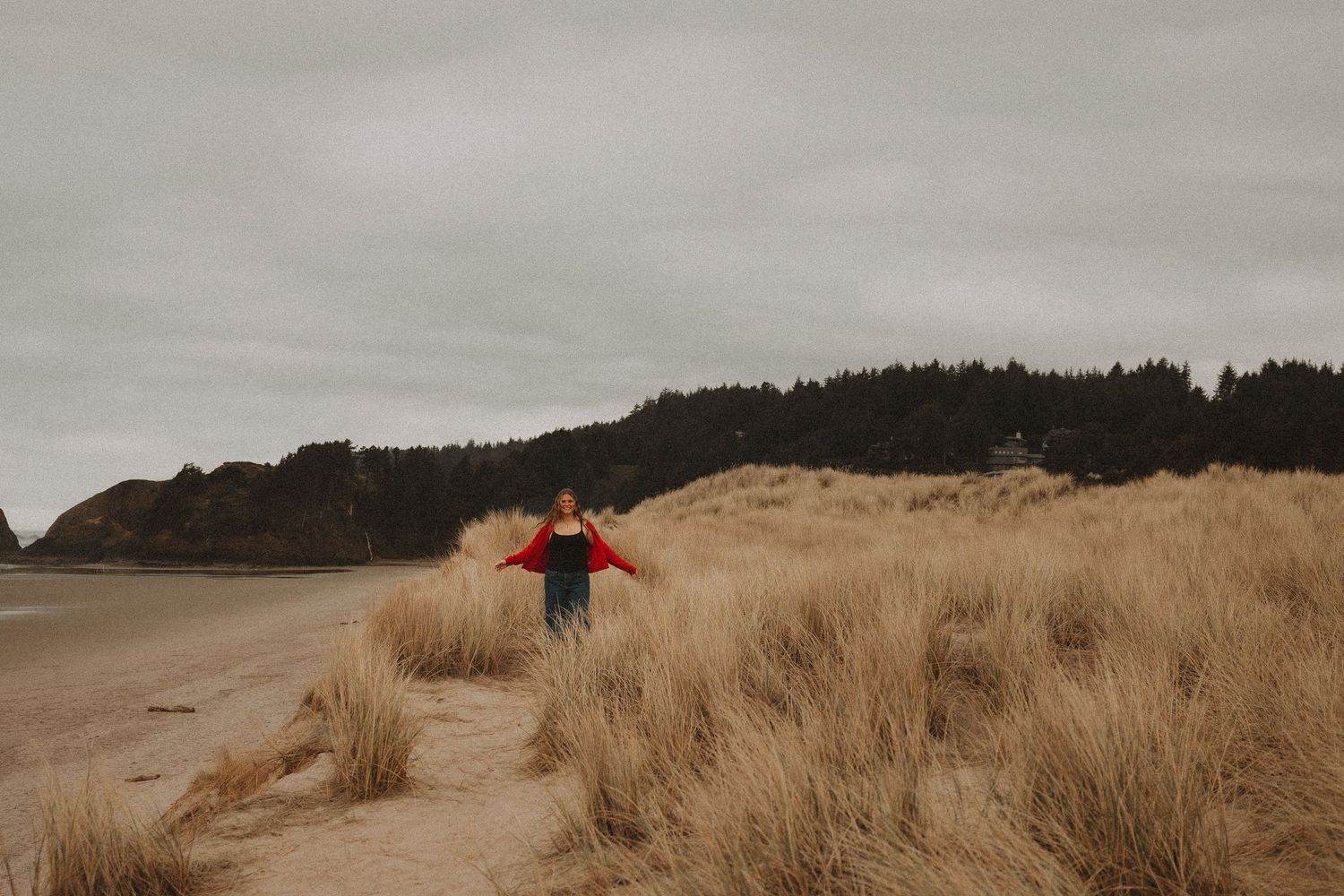A person stands on sandy dunes overlooking a misty Pacific Northwest beach with tall grass and forested cliffs.