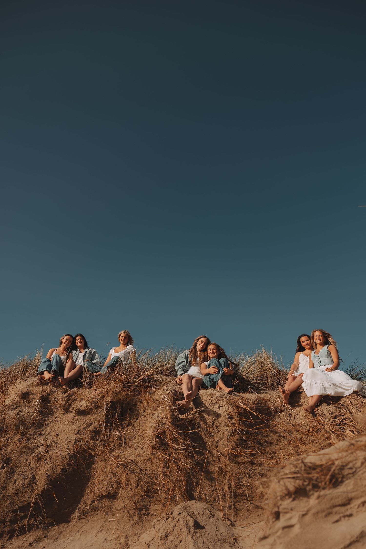 Six young women sit on a sandy grass dune under a deep blue sky at sunset.