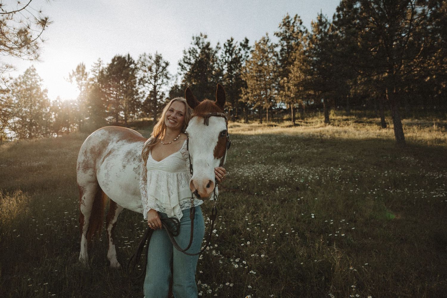 Woman smiling with a brown and white horse in a sunny meadow surrounded by trees.