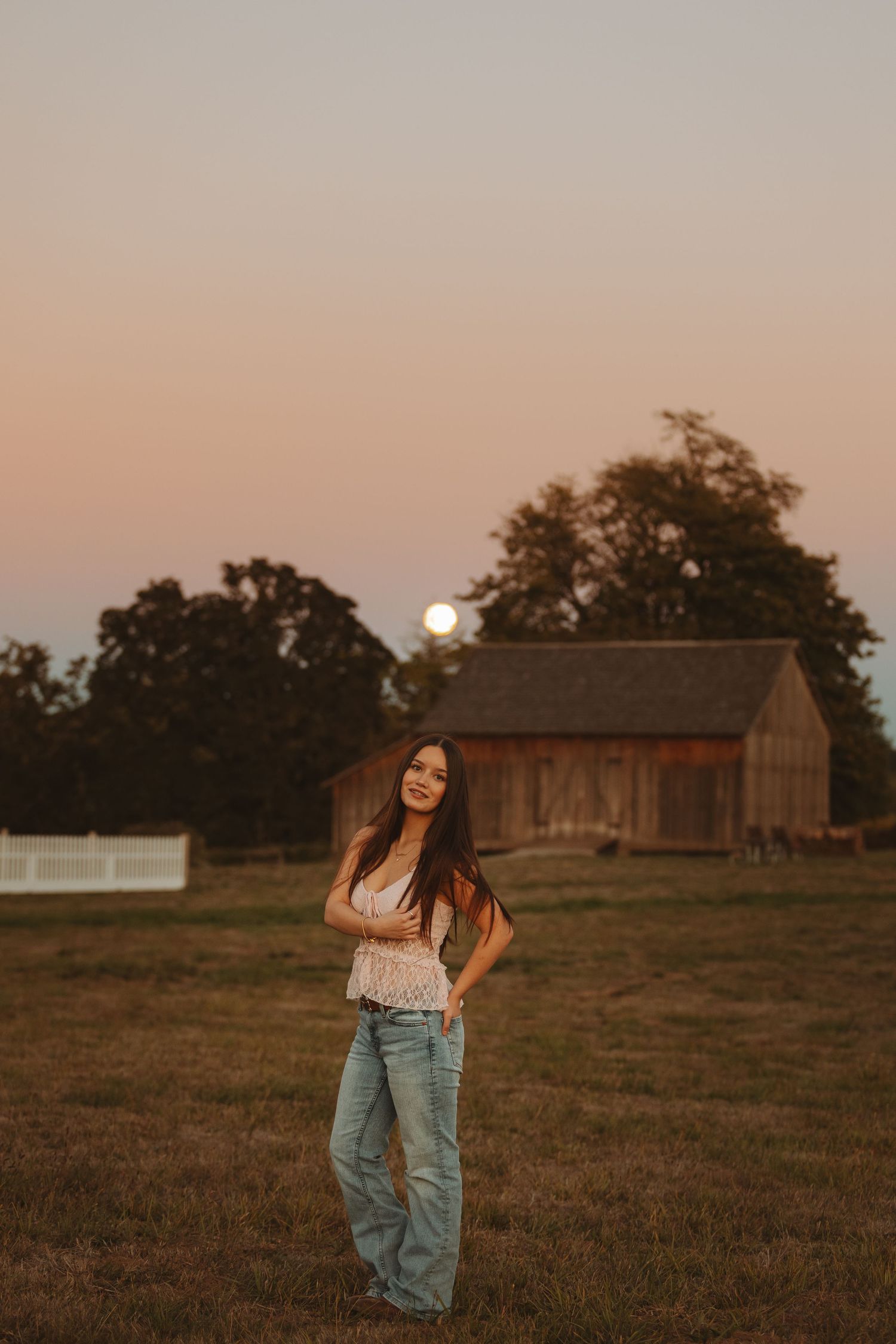 Young woman in jeans and white top poses at golden hour on a farm with a barn and full moon rising.