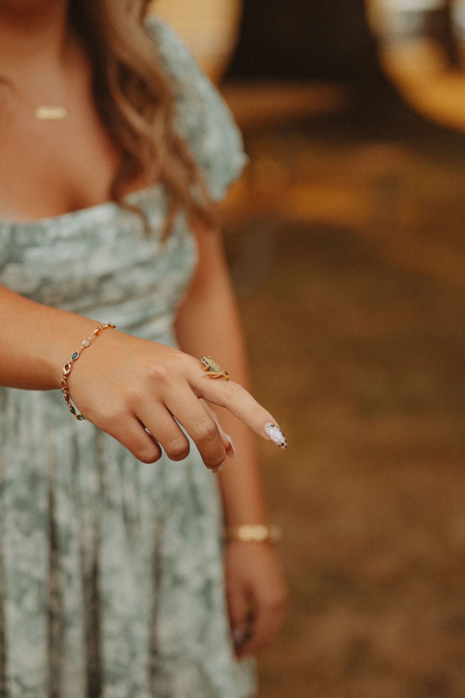 Woman in floral dress wearing gold bracelets and a ring with white nail polish, posed outdoors.