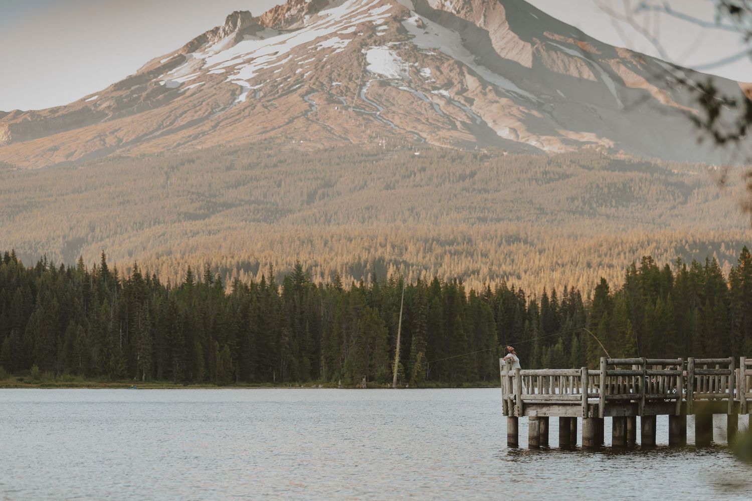 A wooden dock extends over a calm lake with a snow-capped mountain and golden forest in the background.