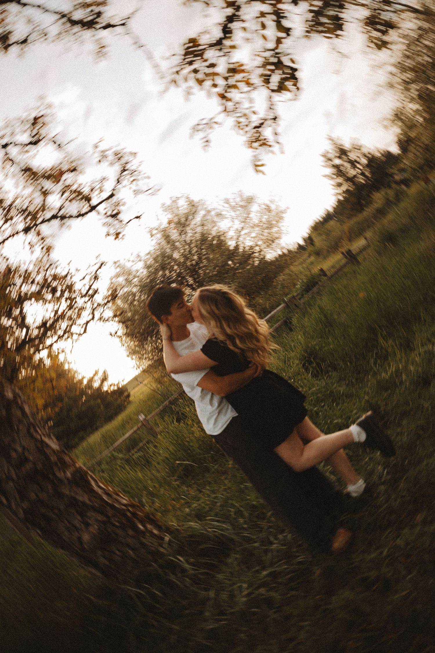 Couple embracing and kissing in a sunny green field at golden hour, captured with a fisheye lens effect.