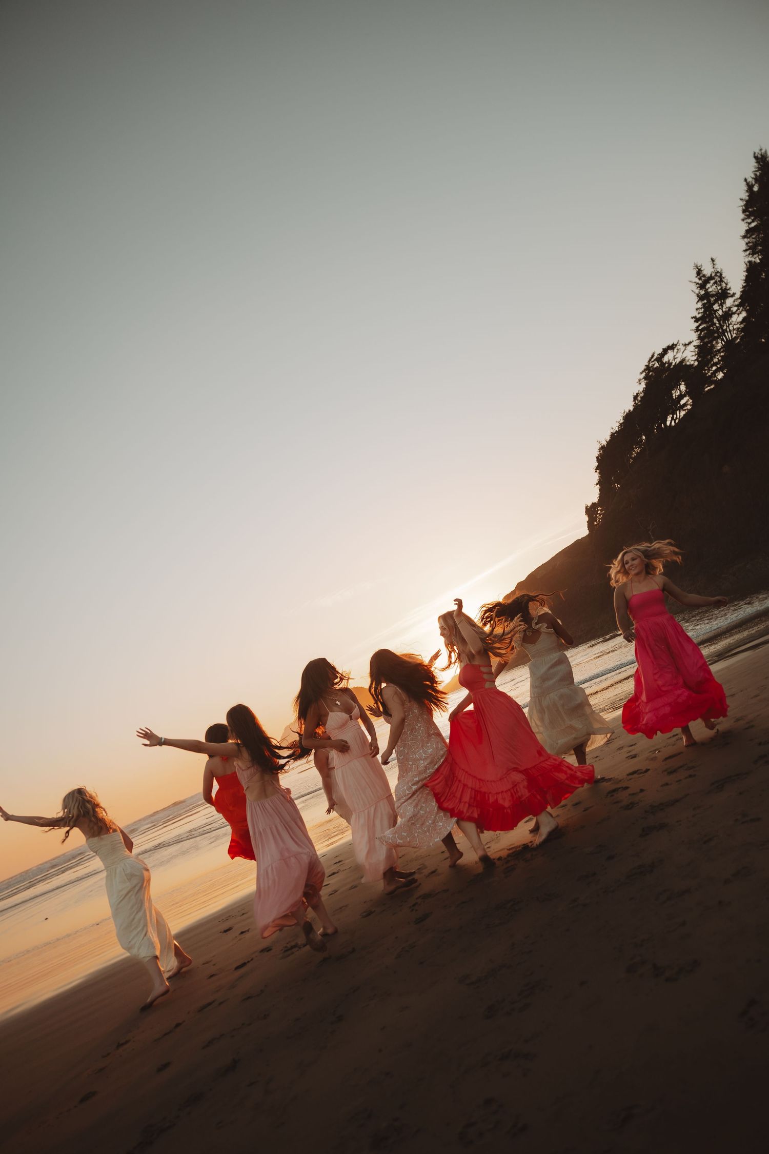 Group of women in flowing red and white dresses dancing on a beach at golden sunset hour.
