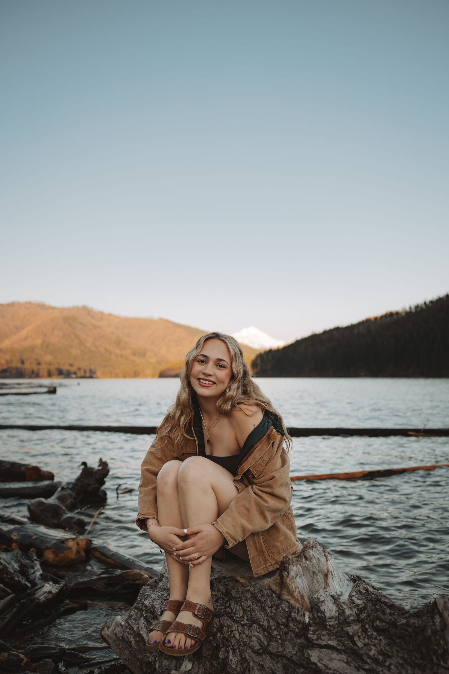 Smiling blonde woman in brown jacket sits on driftwood by a mountain lake at sunset.