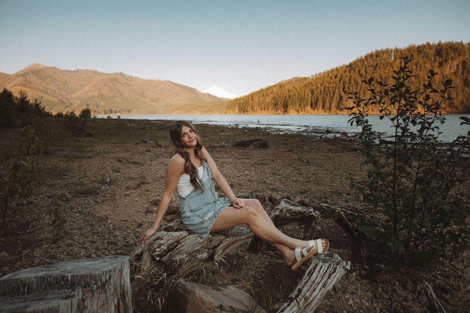 Young woman in denim romper sitting on driftwood logs by a mountain lake at golden hour sunset.