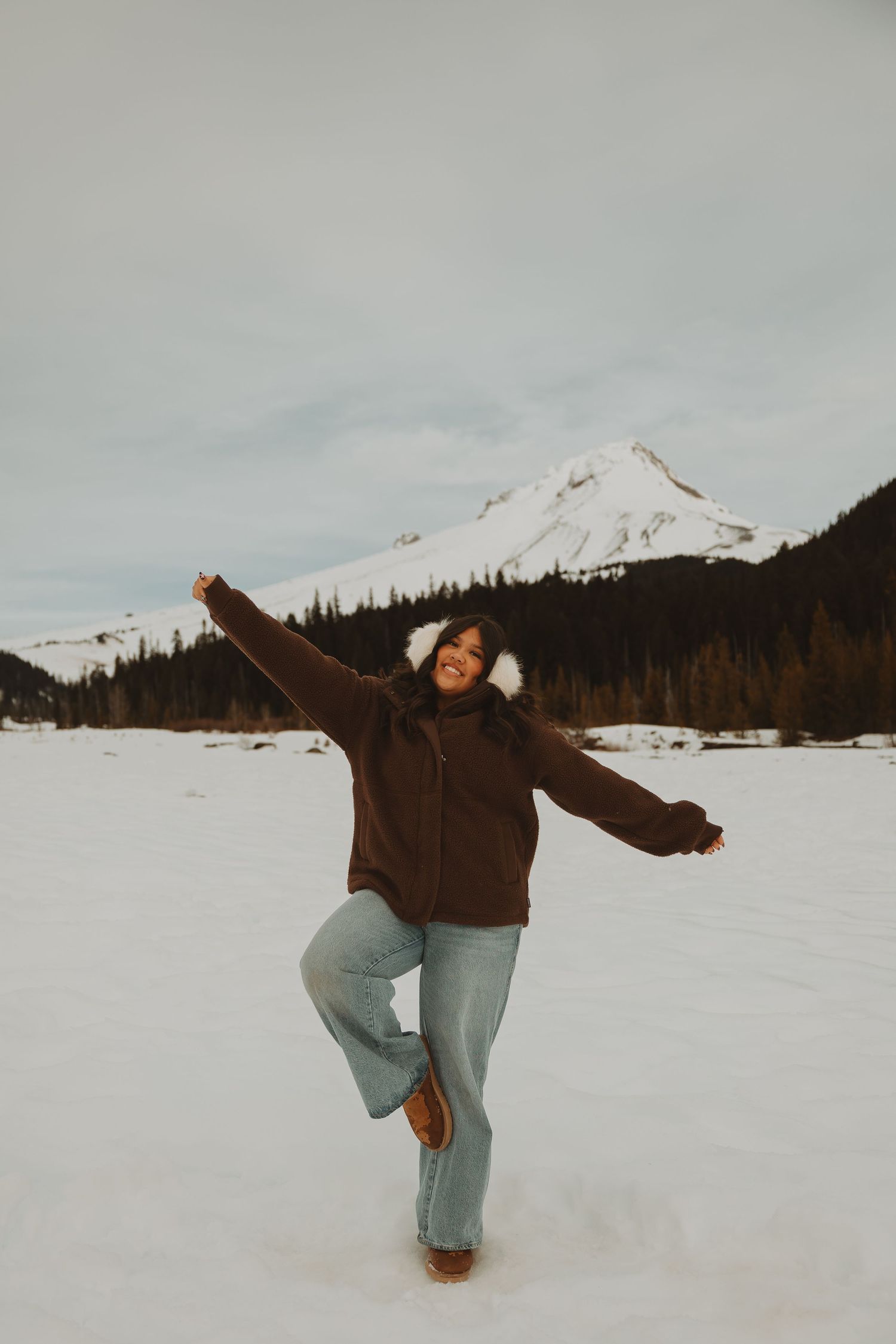 Woman in brown jacket and jeans poses joyfully in snowy mountain landscape with arms outstretched.