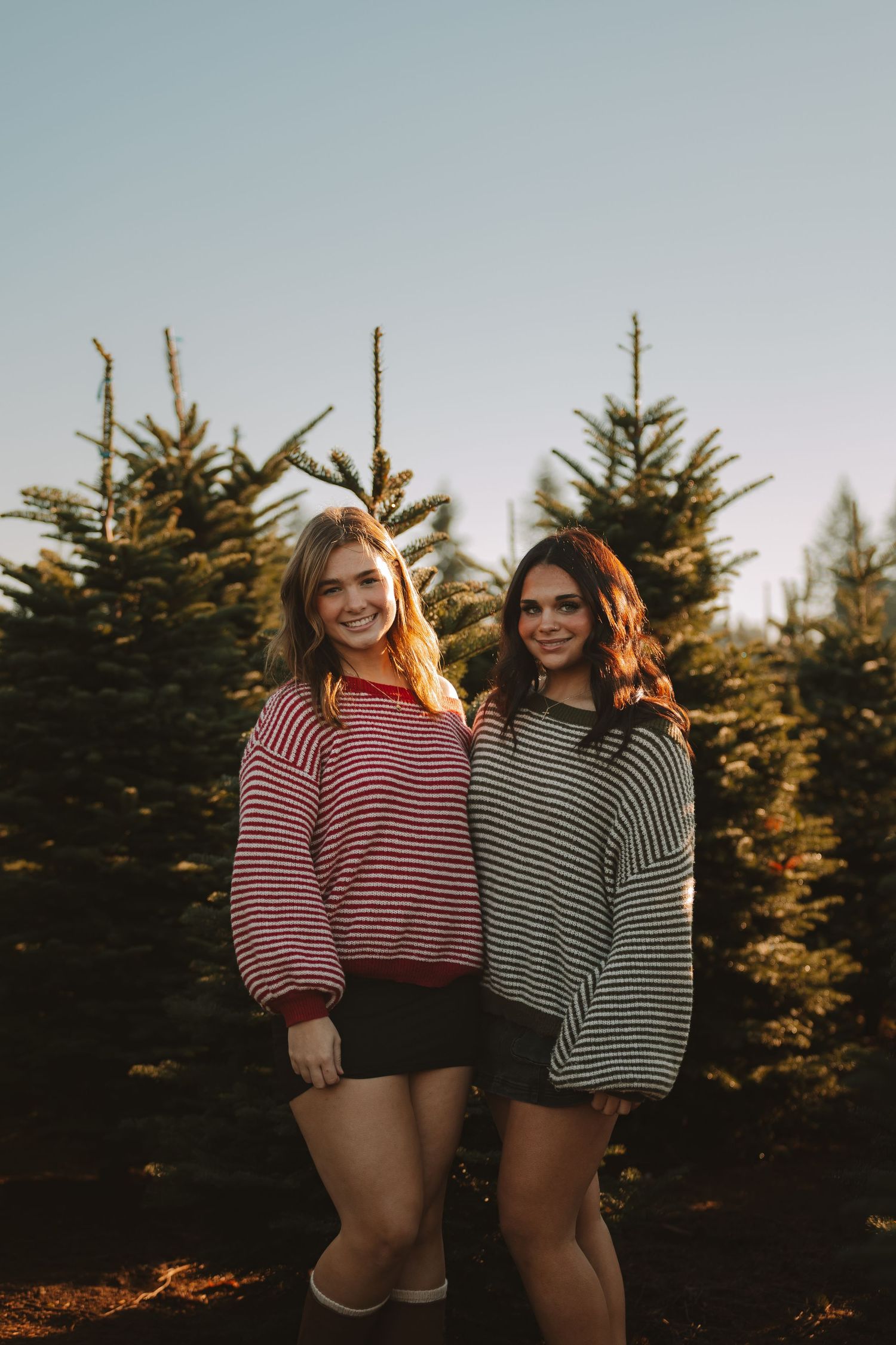 Two people in striped sweaters pose together with evergreen trees in background.