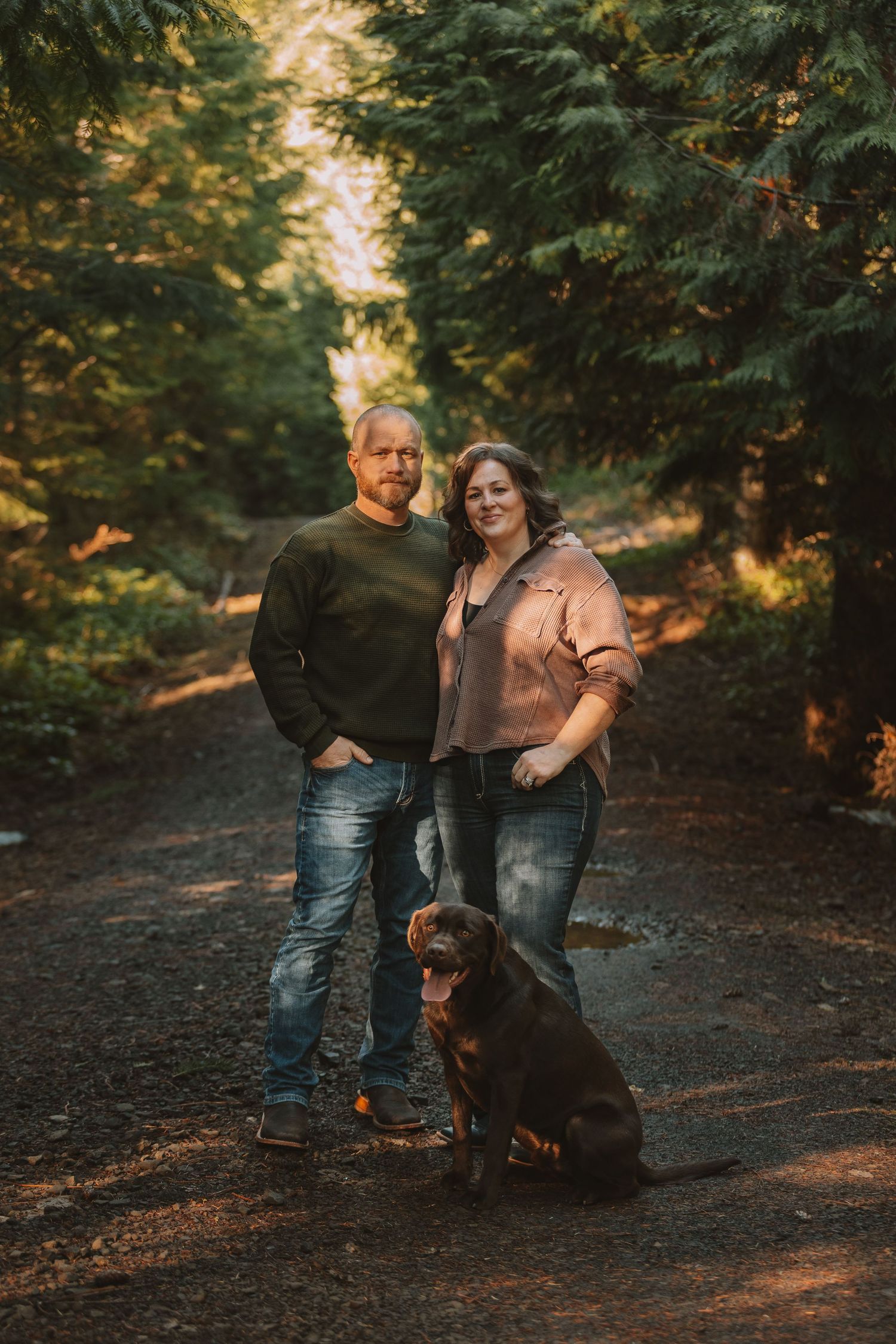 A couple and their chocolate labrador pose together on a wooded path during golden hour sunset.