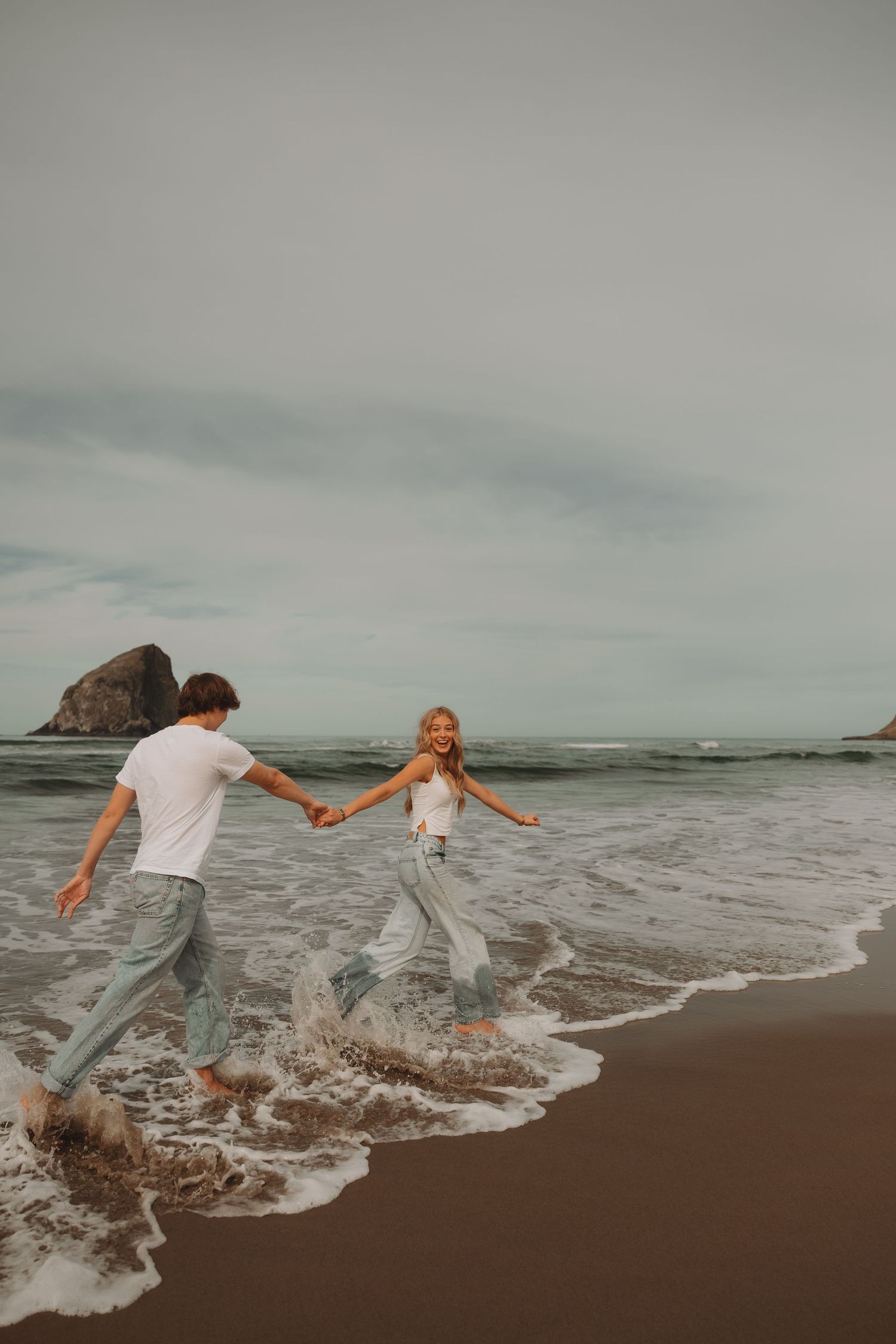 Couple holding hands laughing while running through ocean waves on a sandy beach with rocky formations.