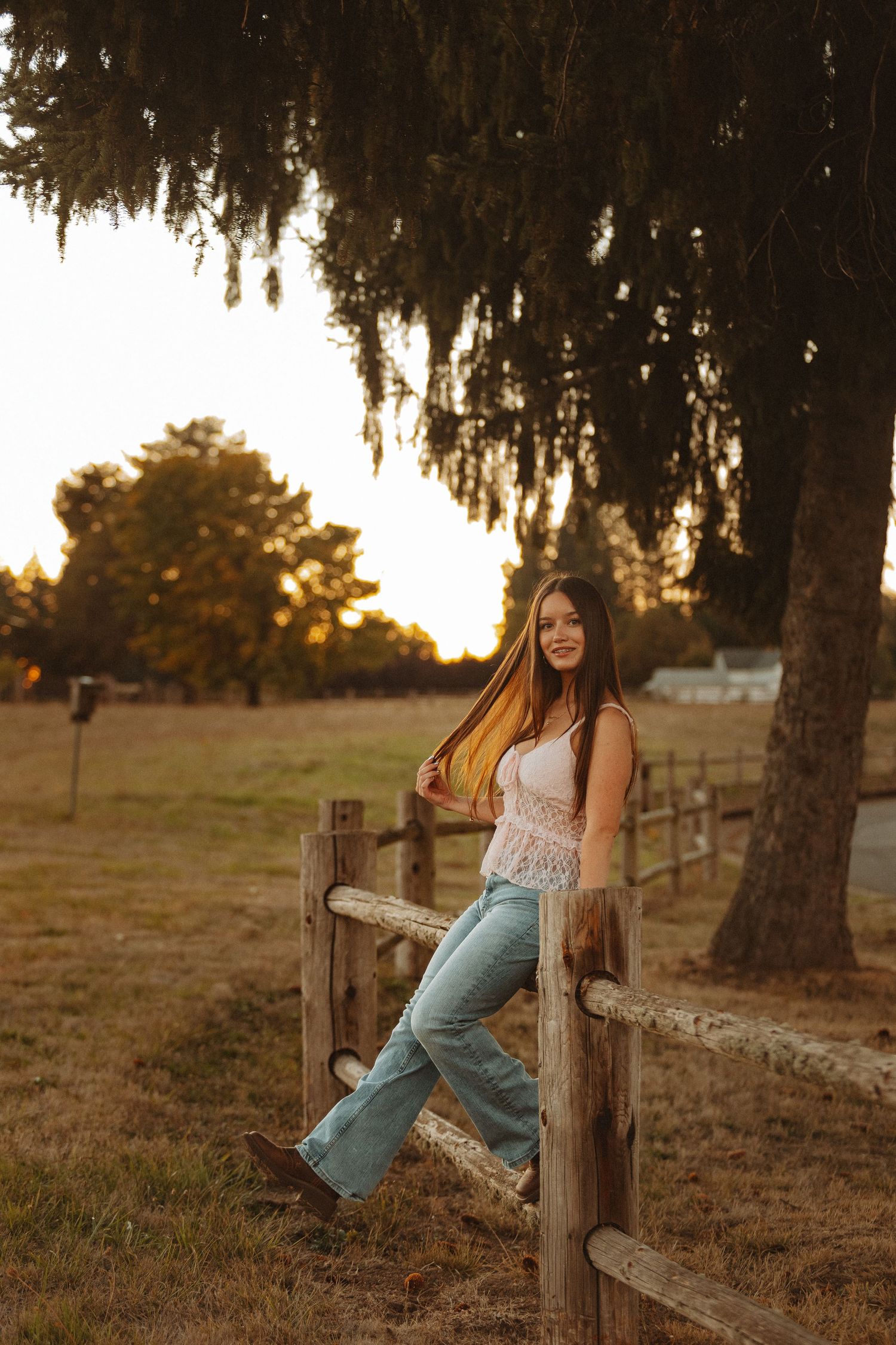 Young woman in flare jeans and lace top leaning on rustic wooden fence at golden hour sunset on a farm.
