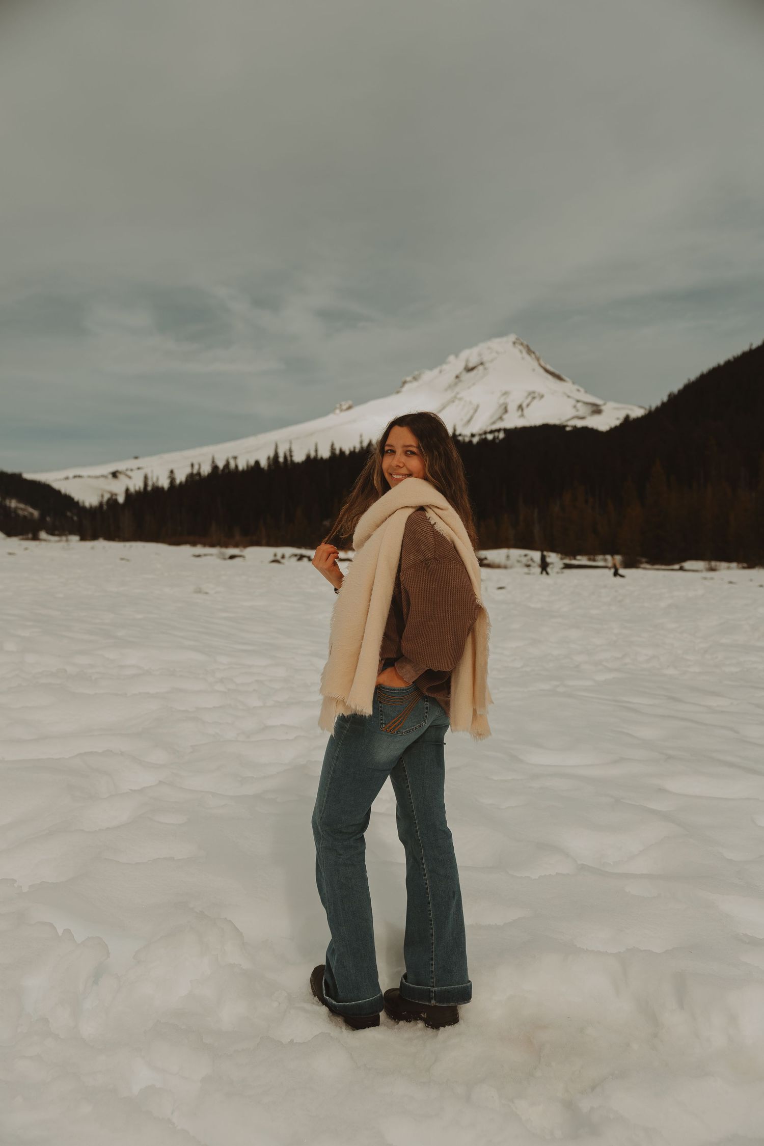 Woman in jeans and cream scarf poses in snowy mountain landscape with snow-capped peak behind her.