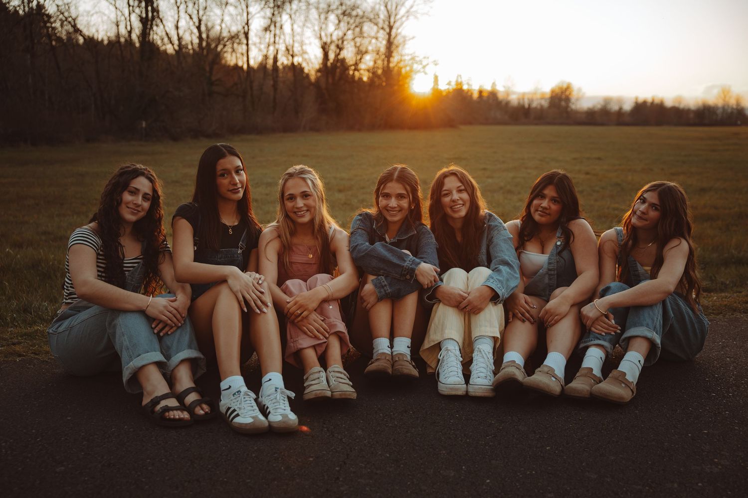 Seven young women sitting together on pavement at golden hour sunset smiling for a group photo outdoors.