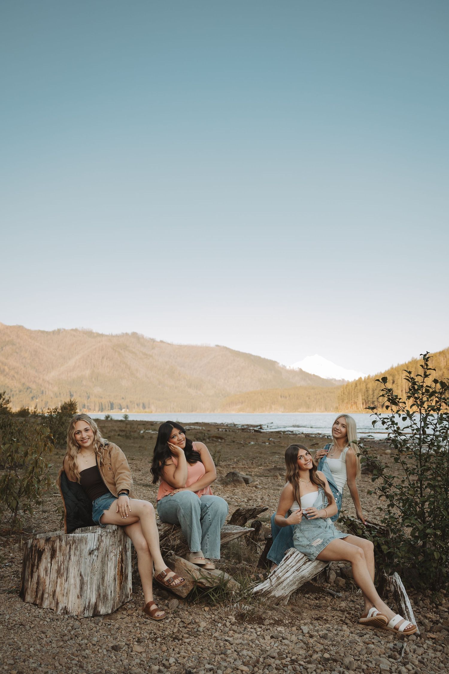 Four friends relax on driftwood logs by a mountain lake during golden hour sunset.