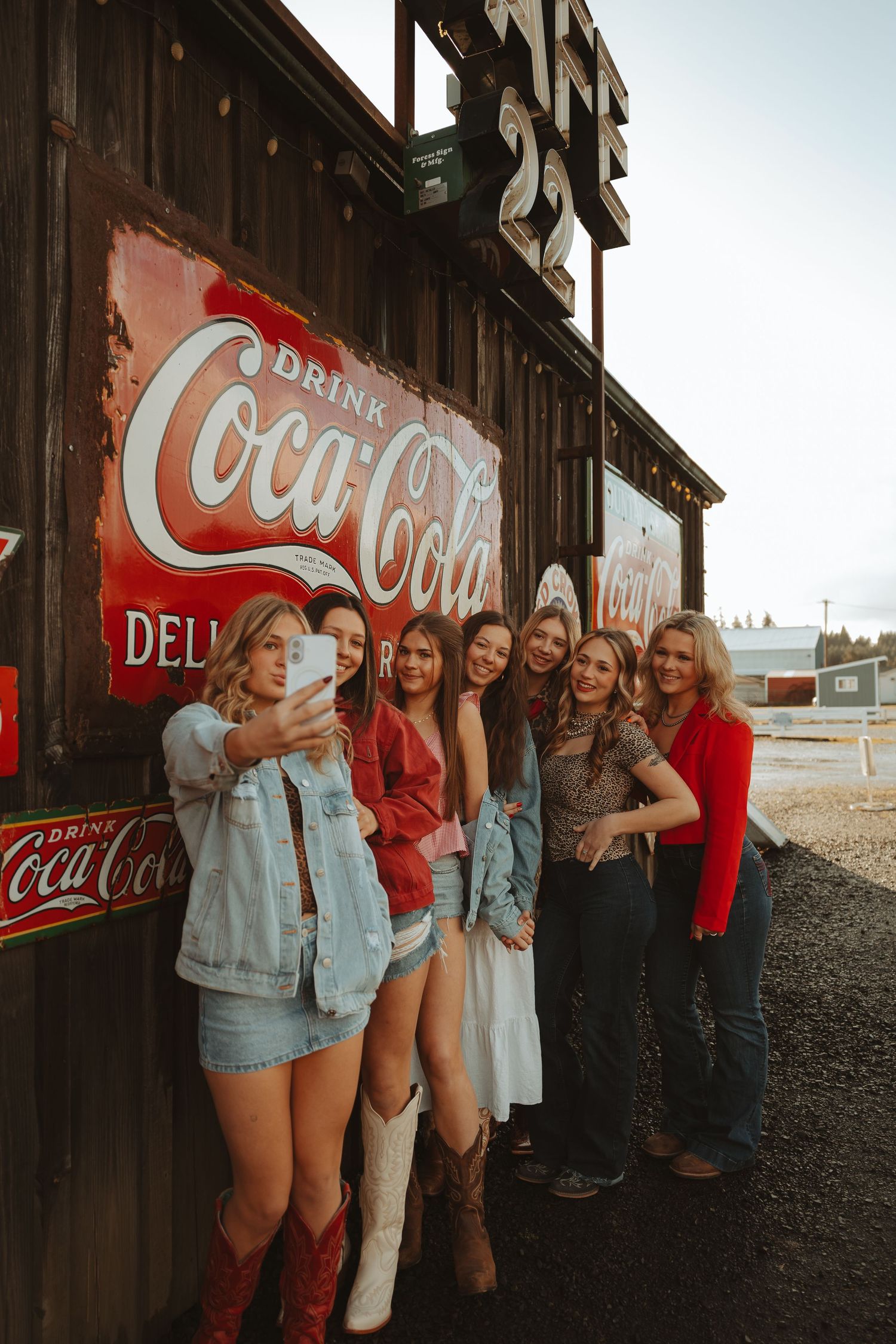 Group of women taking selfie beside vintage Coca-Cola signs on rustic wooden building exterior.