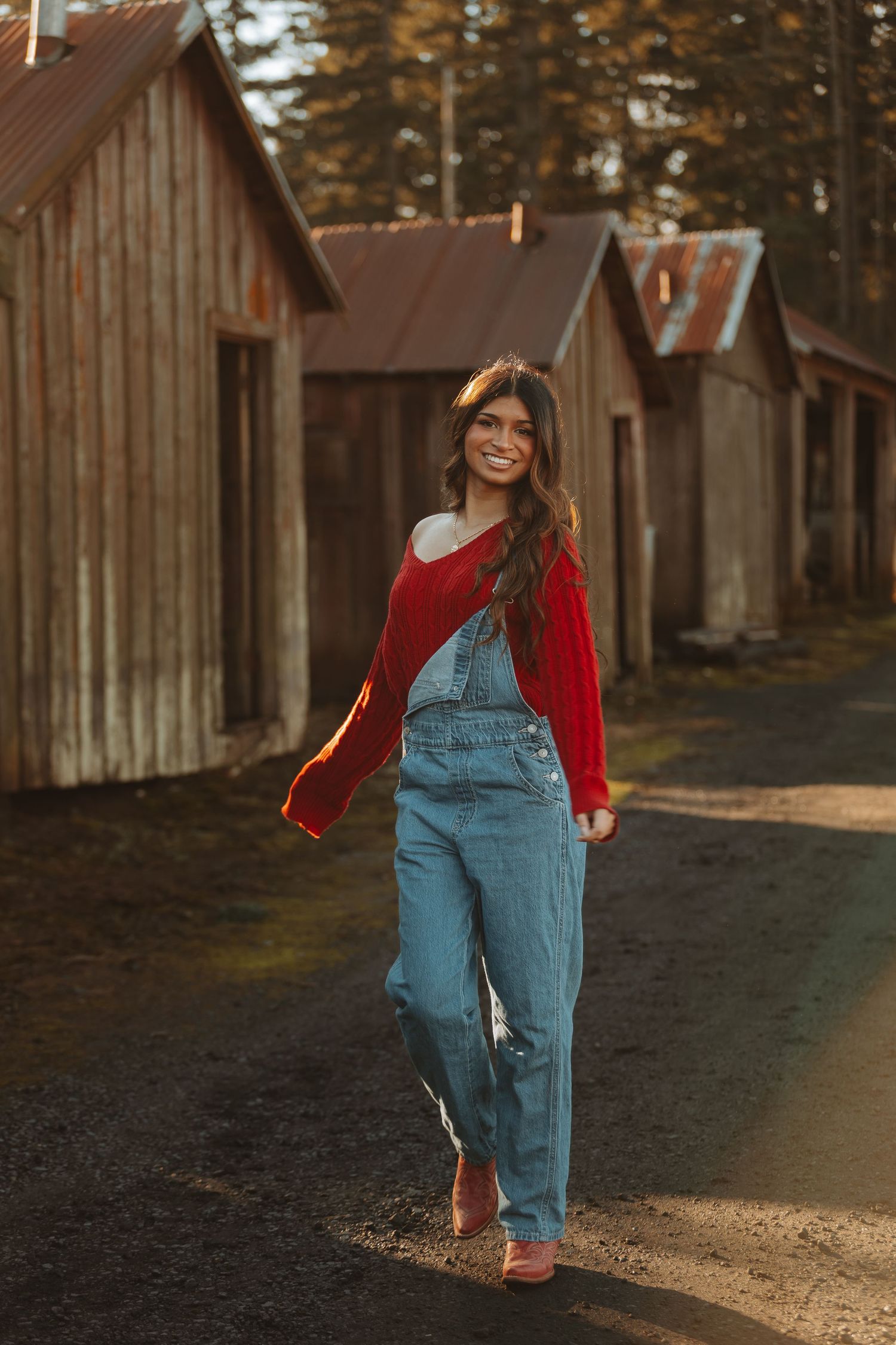 Person in denim overalls and red sweater walks along a path near rustic wooden buildings at sunset.