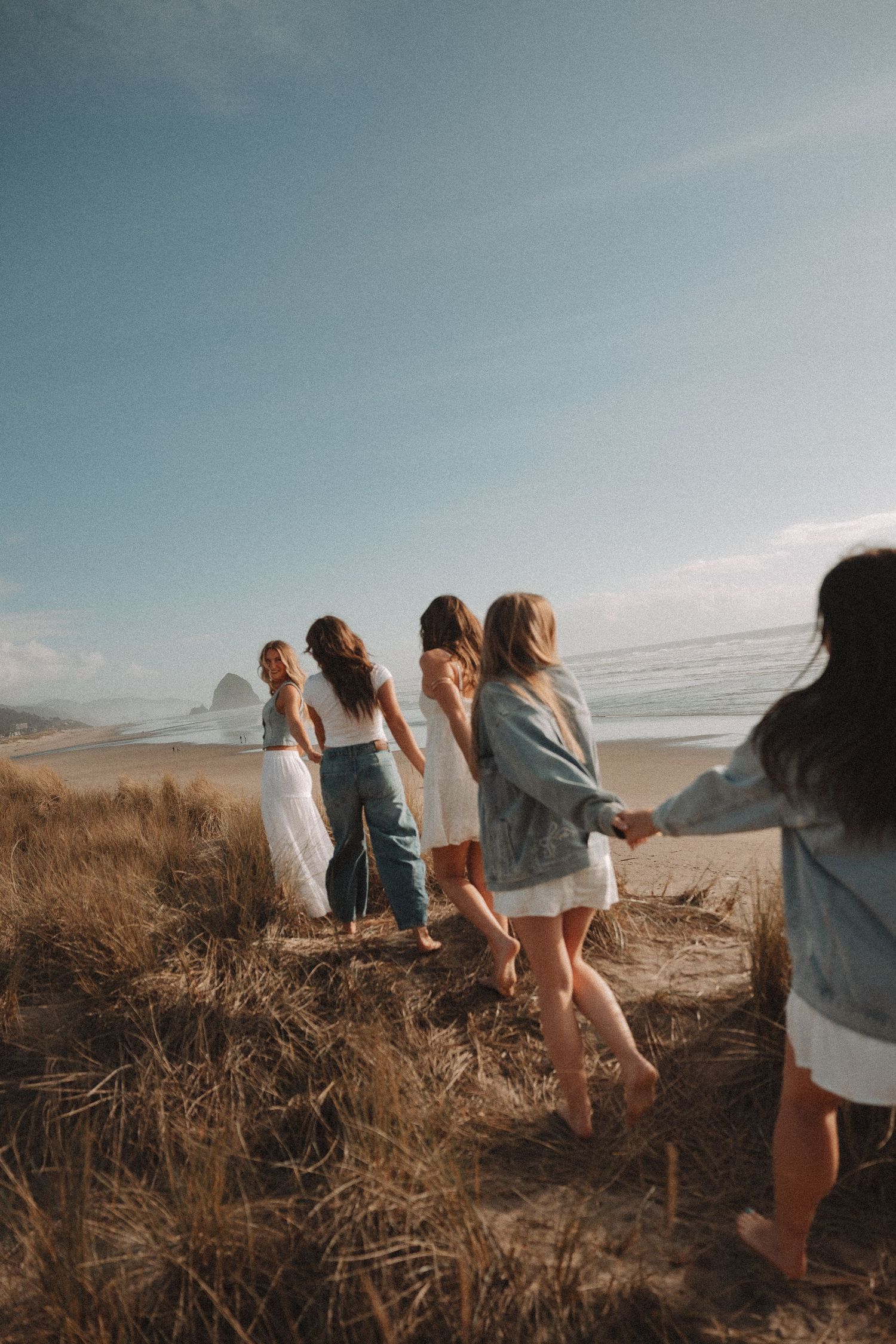 Group of young women holding hands walking through beach dunes toward the ocean.