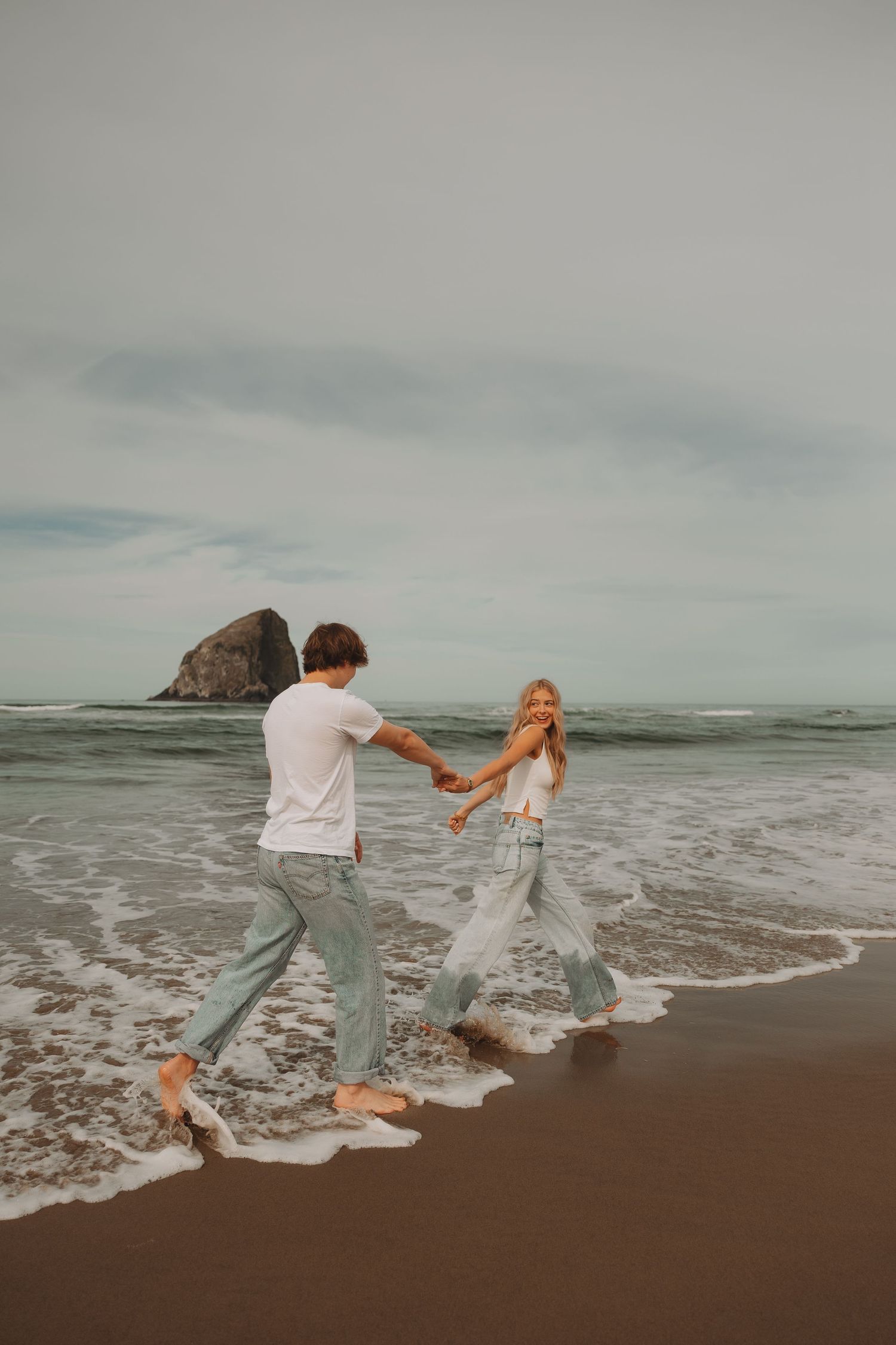 Couple laughs and runs through ocean waves on sandy beach with rock formation behind.