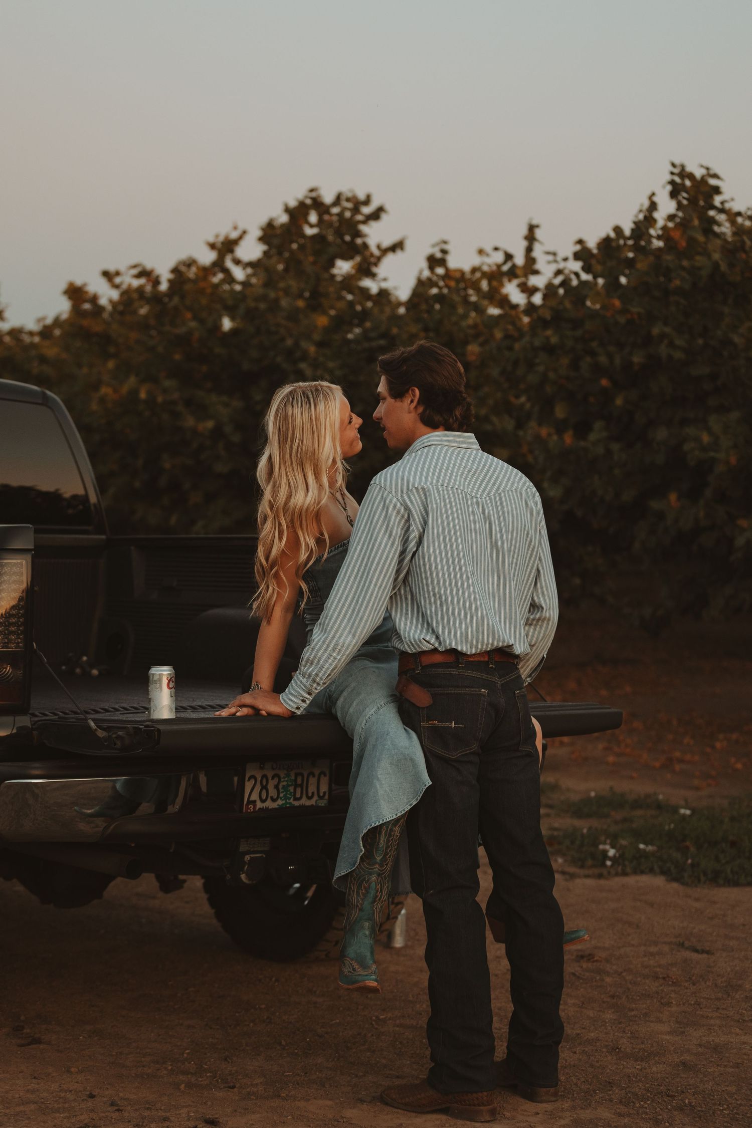Couple leaning in for a kiss on a truck tailgate at sunset with vineyard in background.