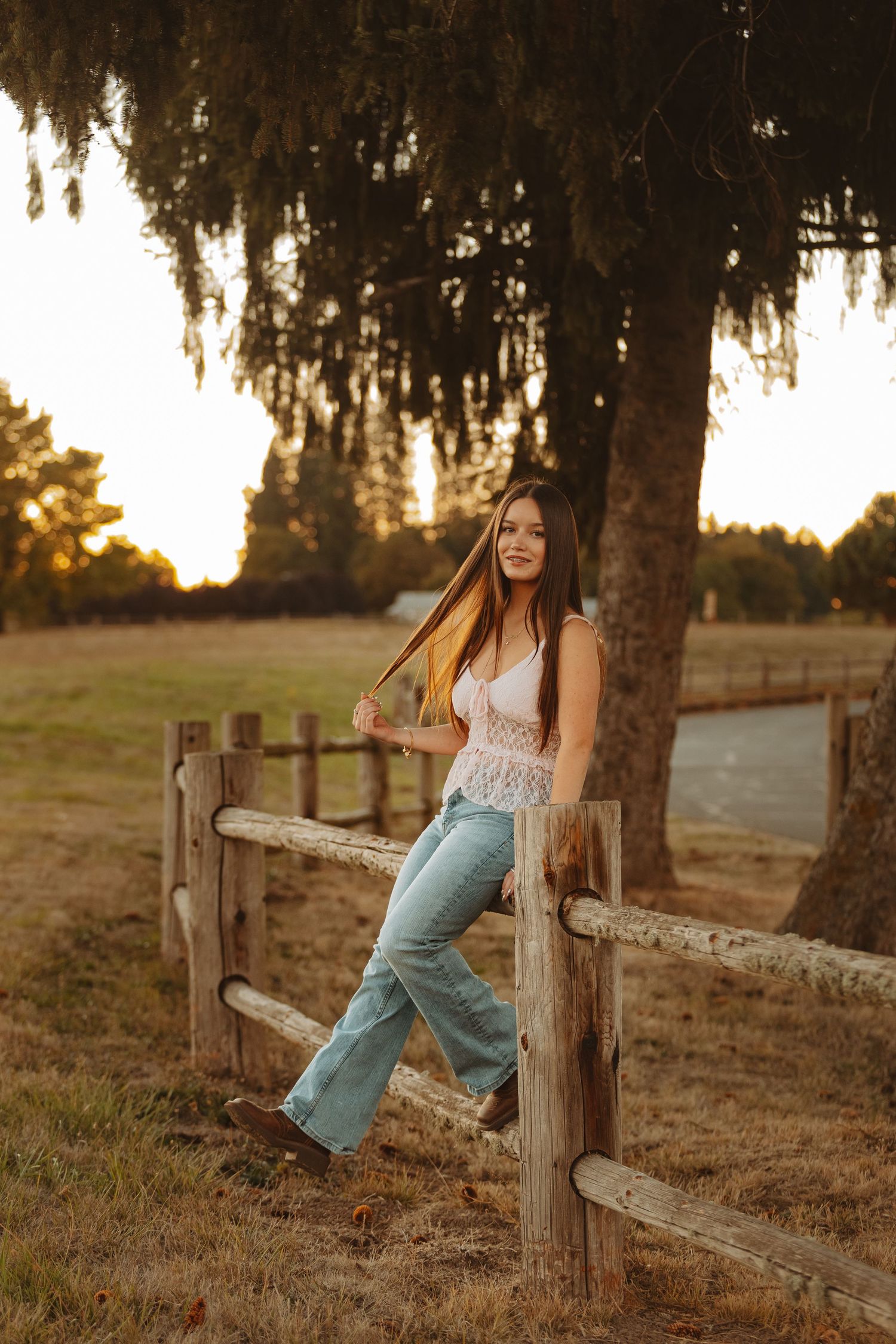 Young woman in white lace top and flare jeans leaning on rustic wooden fence at golden sunset on a farm.