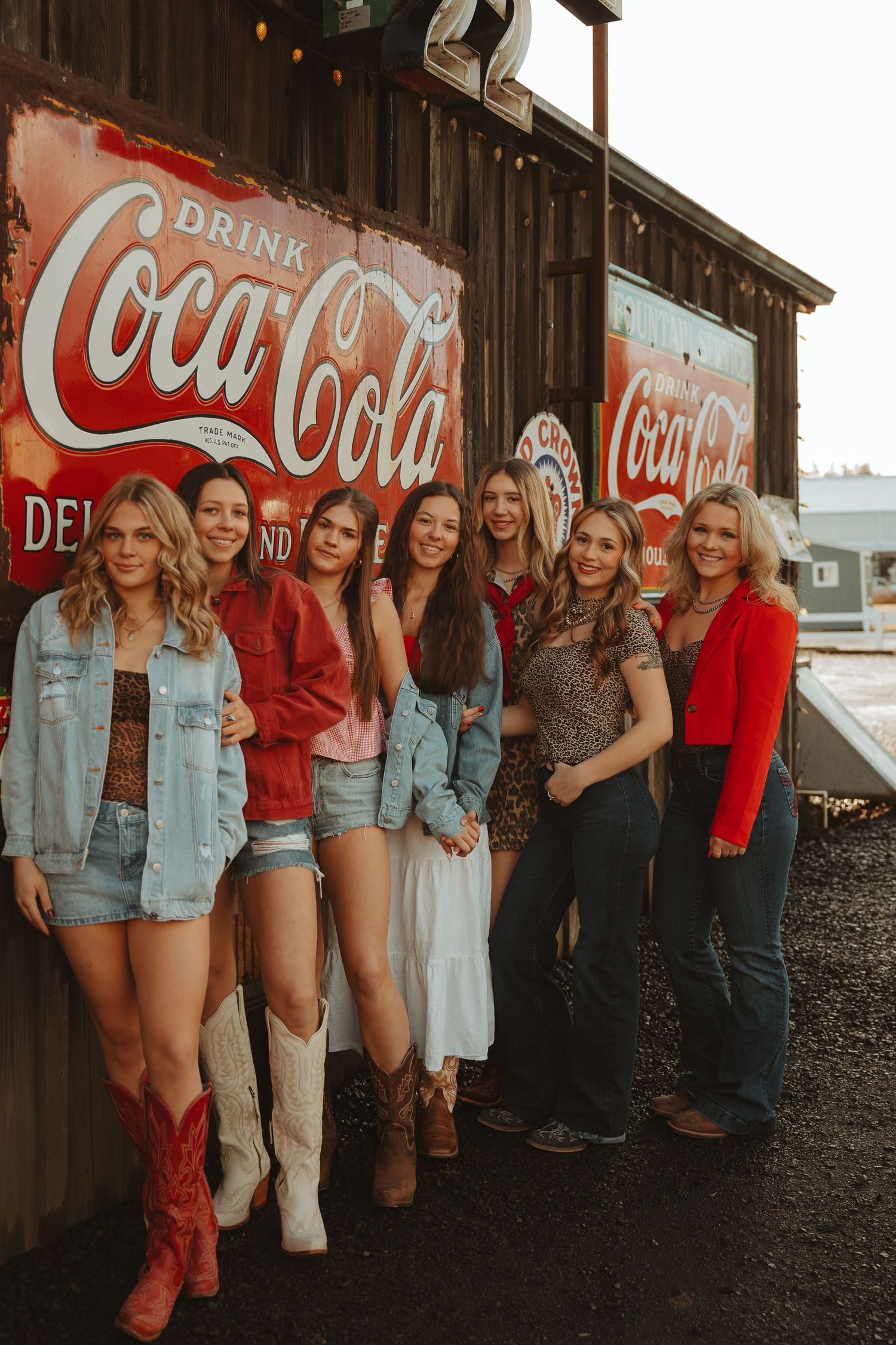 Group of seven women posing in front of vintage Coca-Cola signs, wearing western outfits with cowboy boots.