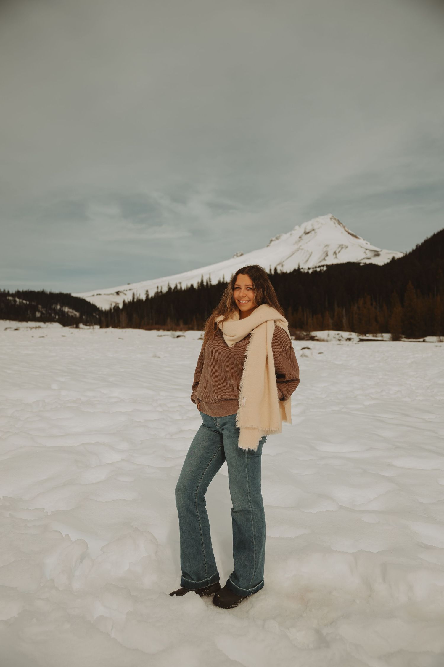 Smiling woman in brown sweater cream scarf and flare jeans stands on snowy field near mountain.
