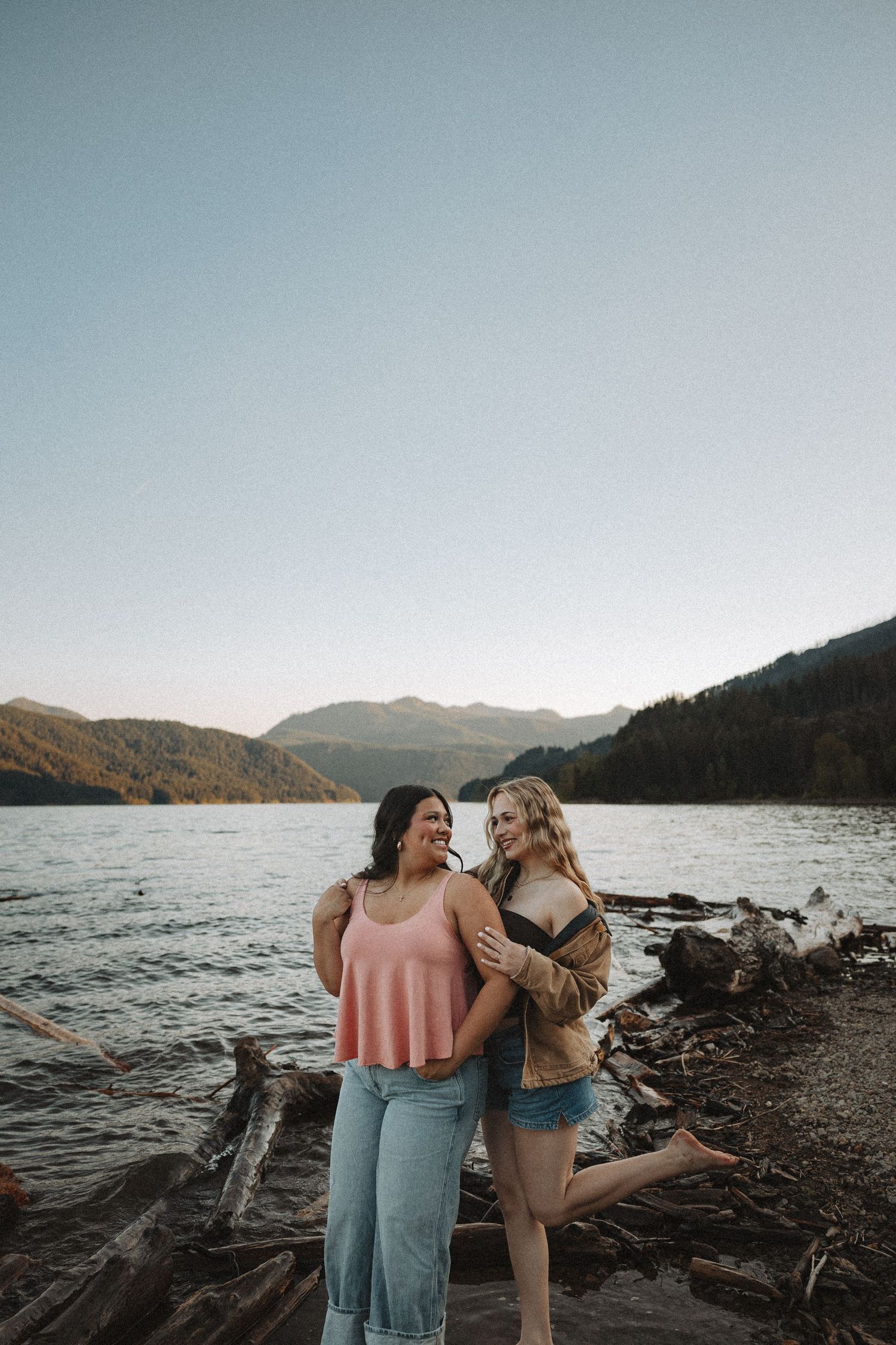 Two young women laughing and embracing on a rocky lakeshore with mountain backdrop at sunset.