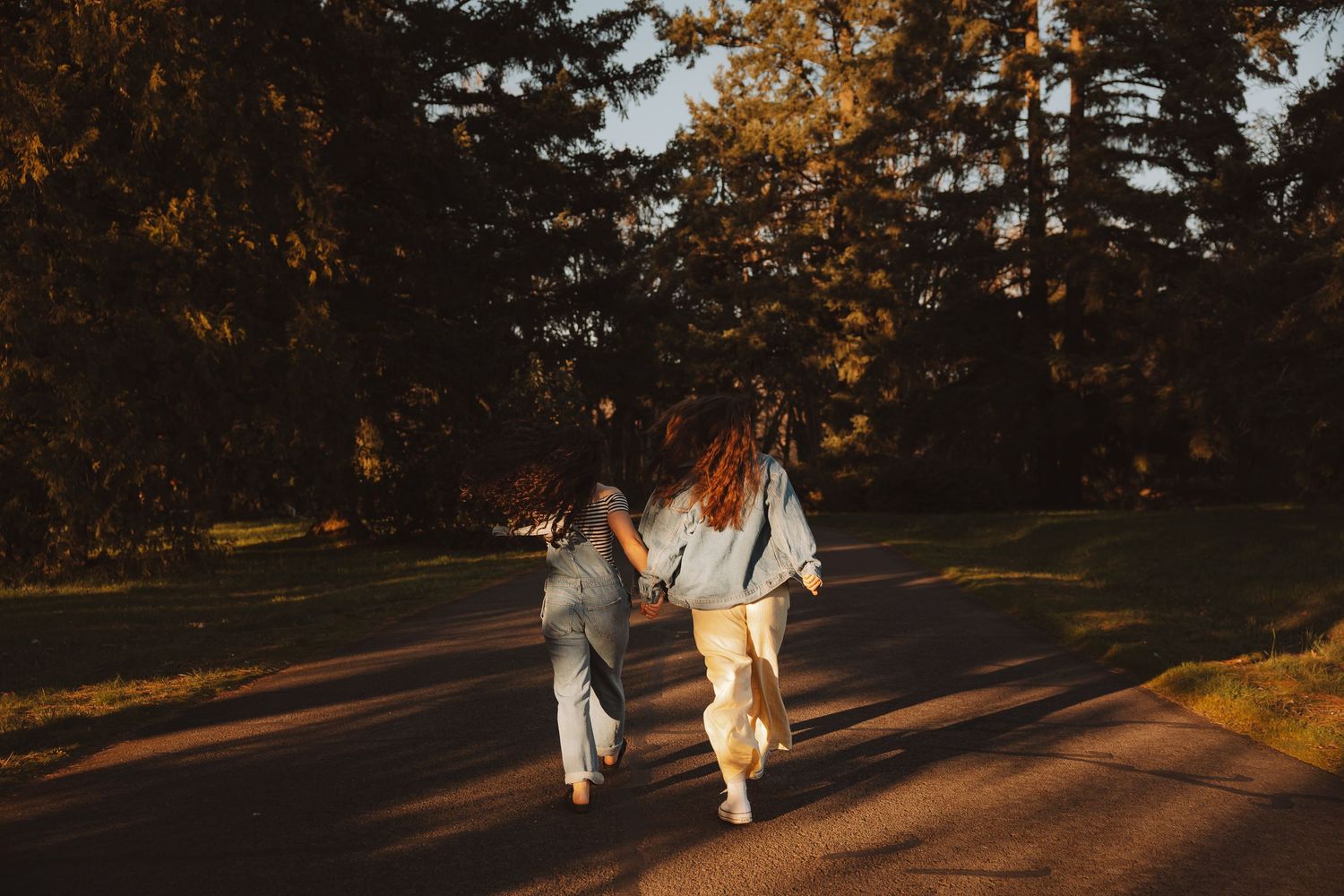 Two friends holding hands running down a tree-lined road during golden hour.