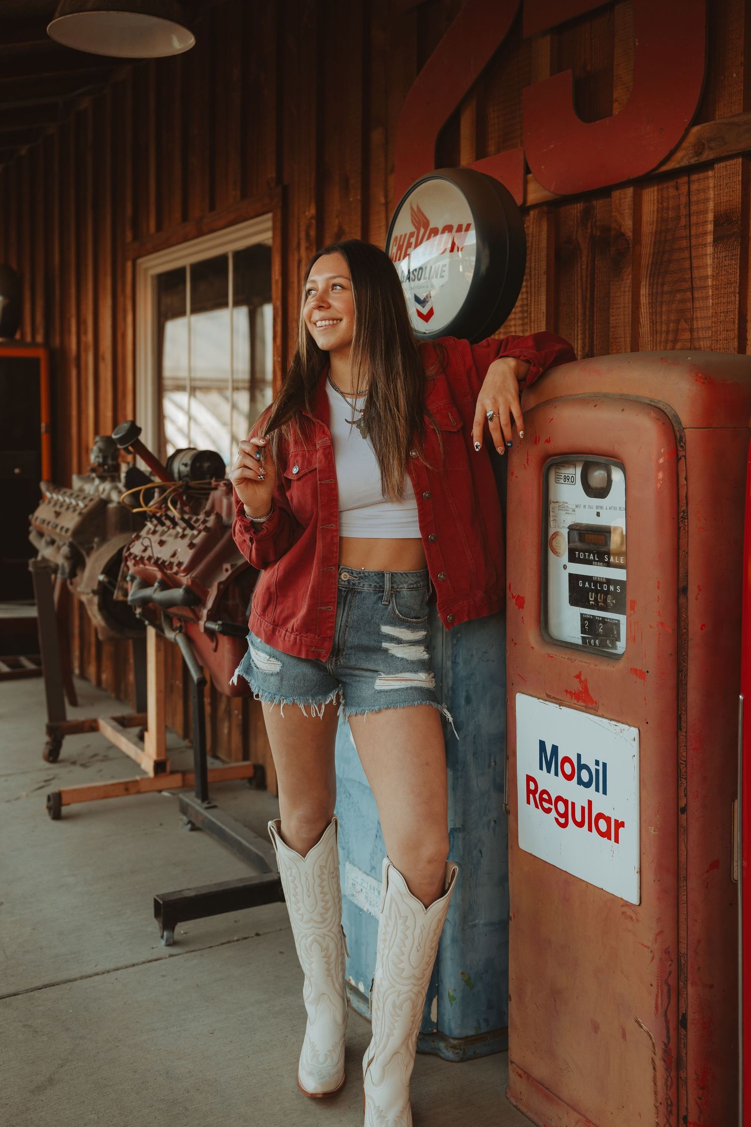 Woman in red jacket, denim shorts, and white cowboy boots leaning against a vintage Mobil Regular gas pump in a rustic garage.