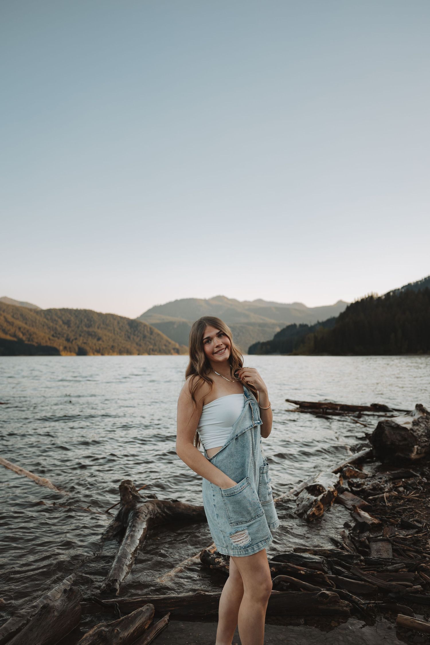 Young woman in denim overalls and white top smiling by a mountain lake with driftwood at sunset.
