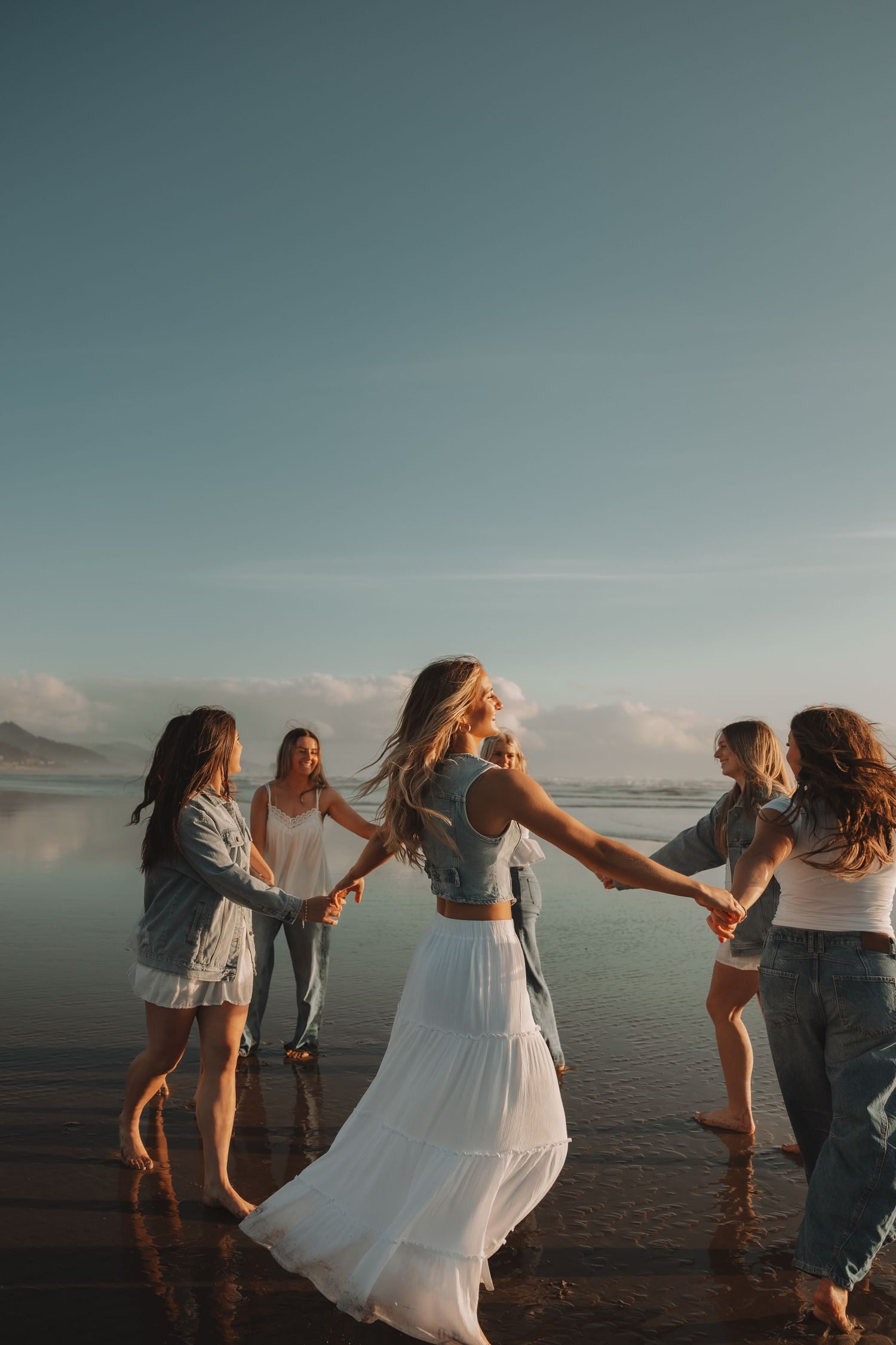 Group of women hold hands dancing in a circle on a beach at golden hour sunset.