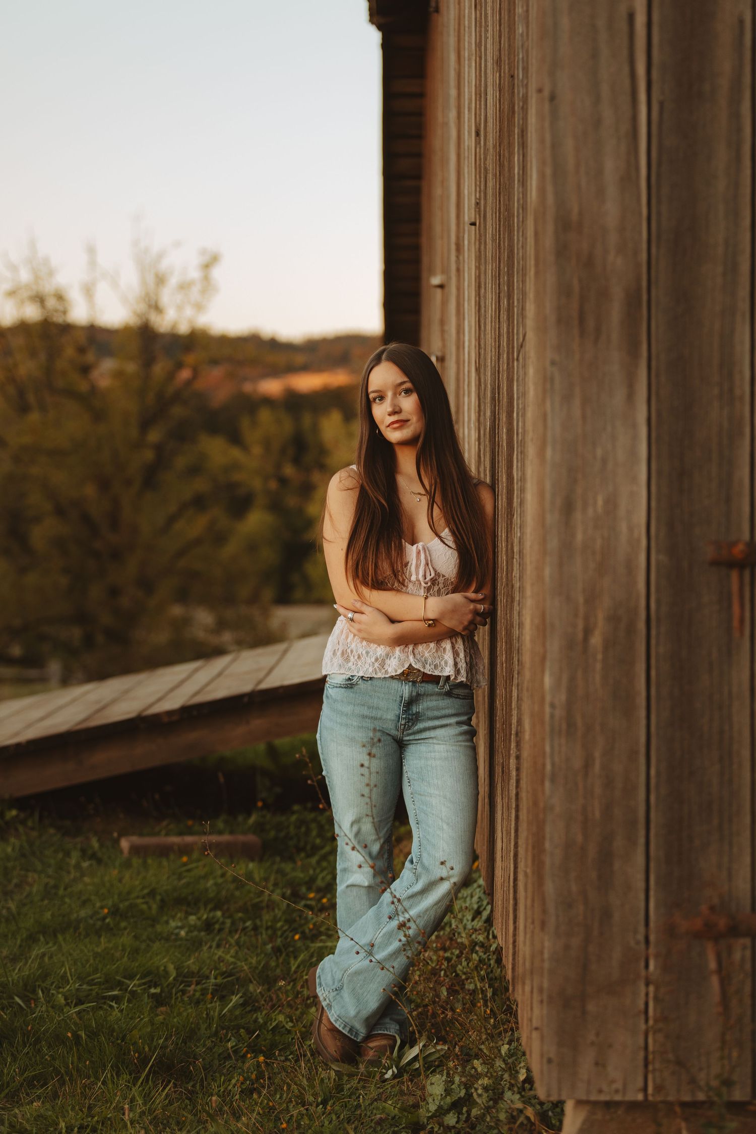 Young woman with long hair leaning against rustic wooden barn wall at golden hour, wearing white top and jeans.