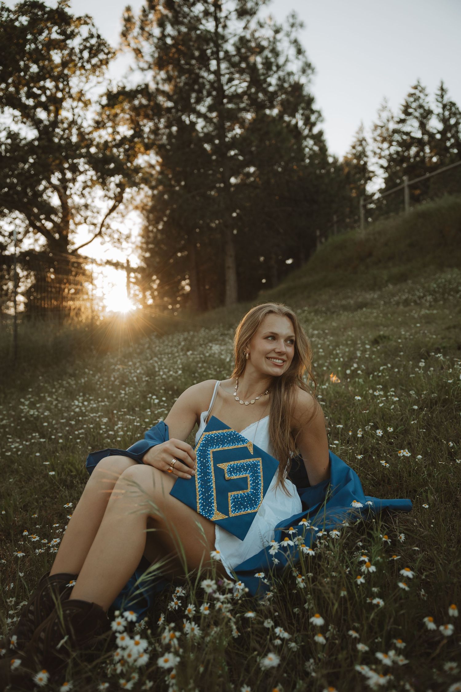 Smiling young woman sits in daisy field at sunset, holding blue graduation items, wearing white dress and brown boots.