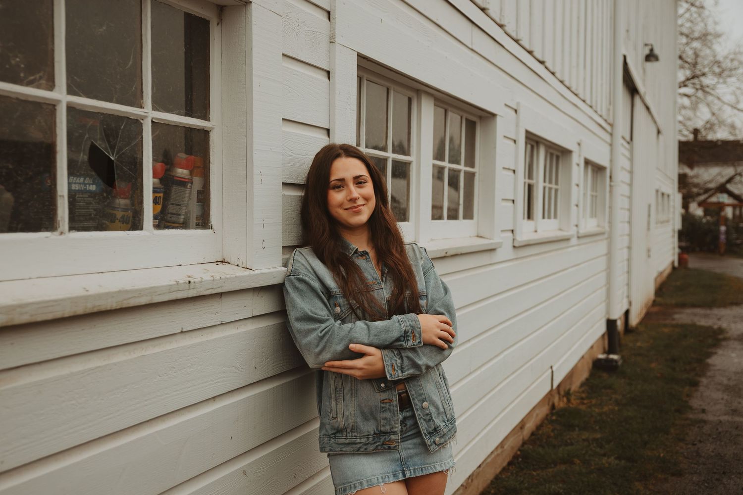 A woman in a denim jacket poses casually against a white building exterior in natural lighting.