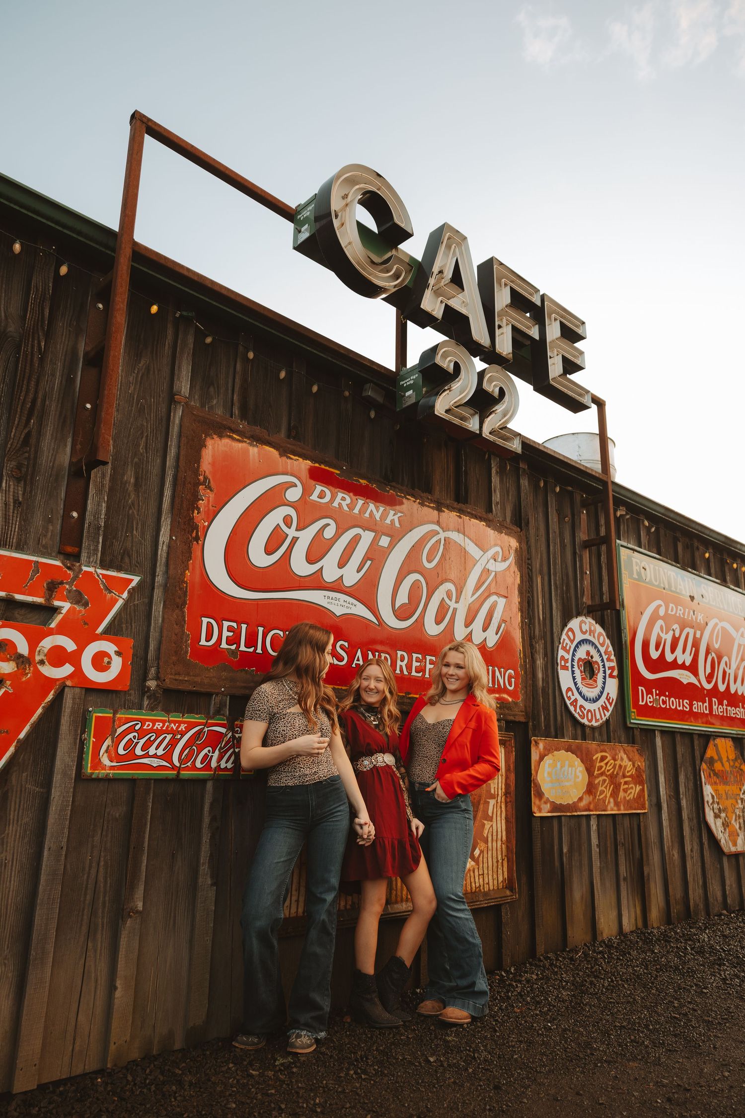 Three women pose under a Cafe 22 neon sign surrounded by vintage Coca-Cola advertisements on a wooden wall.