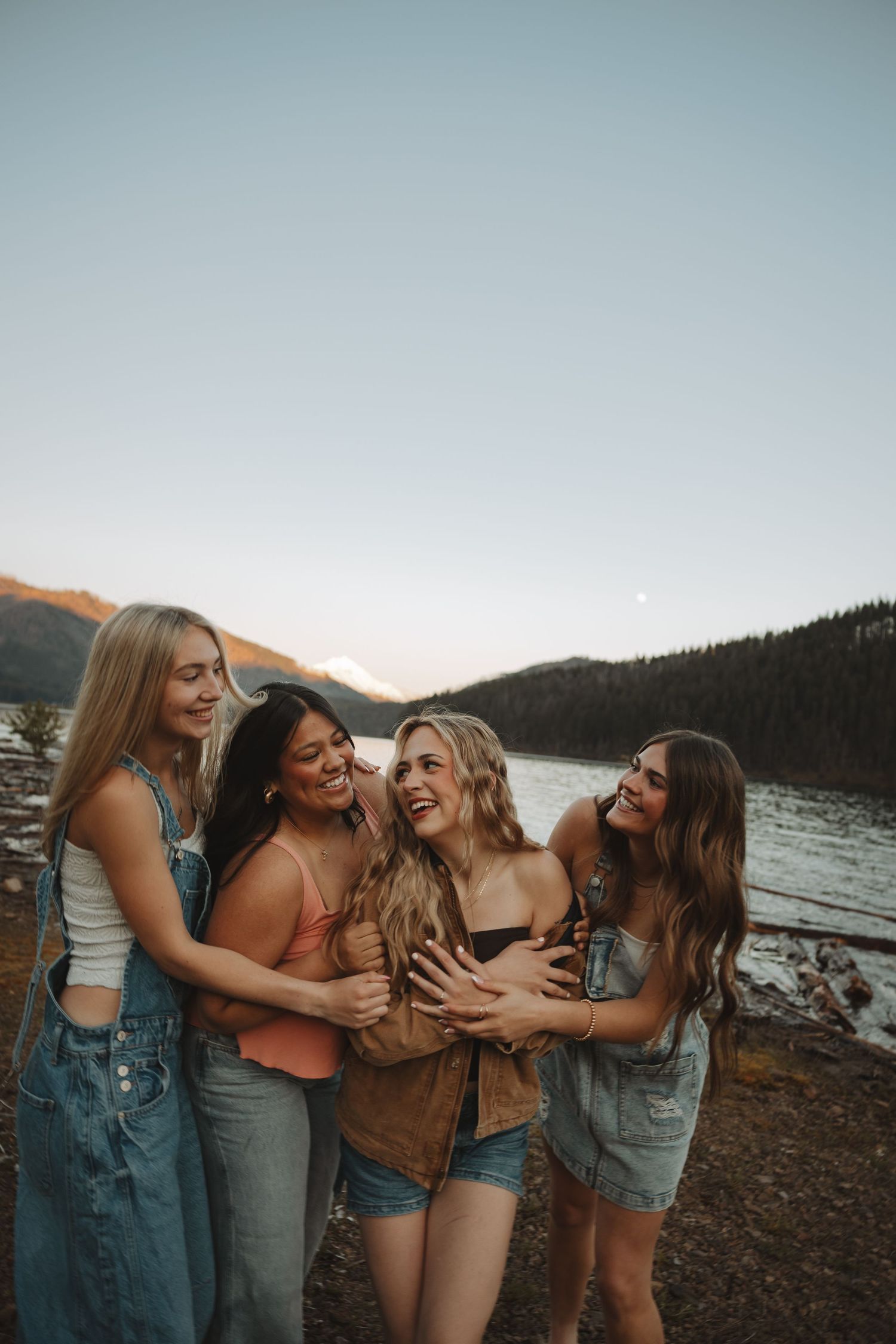Four young women smiling and embracing by a mountain lake at dusk.