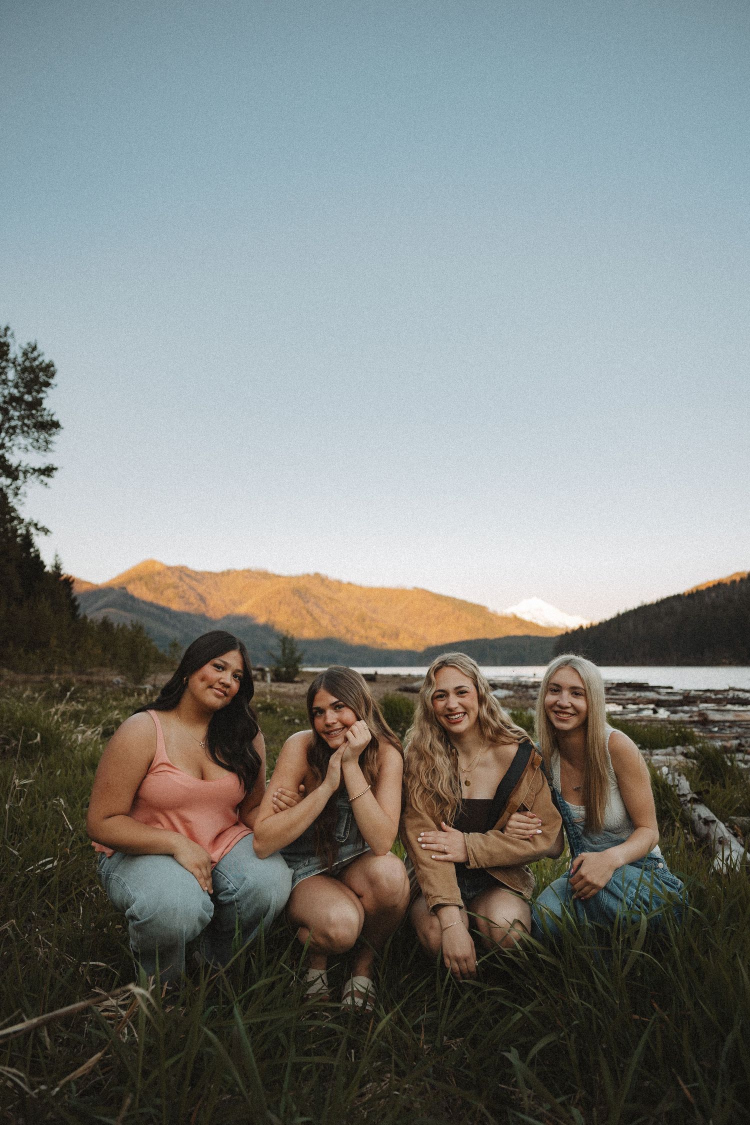 Four smiling young women posing together in a mountain lakeside meadow at sunset.