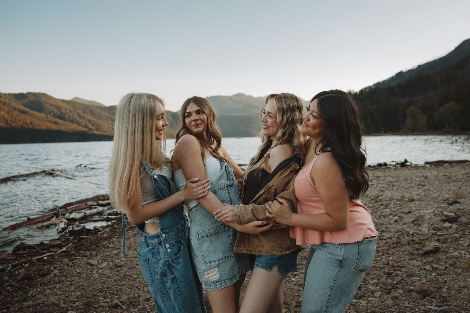 Four young women laughing and embracing near a mountain lake at sunset.