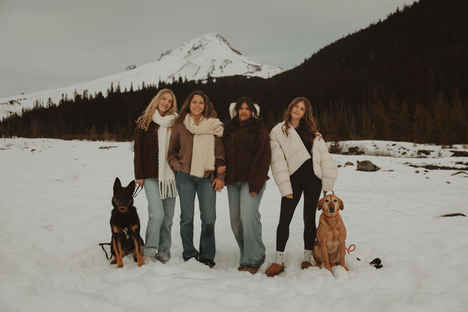 Four women posing with two dogs in a snowy mountain landscape wearing winter outfits.