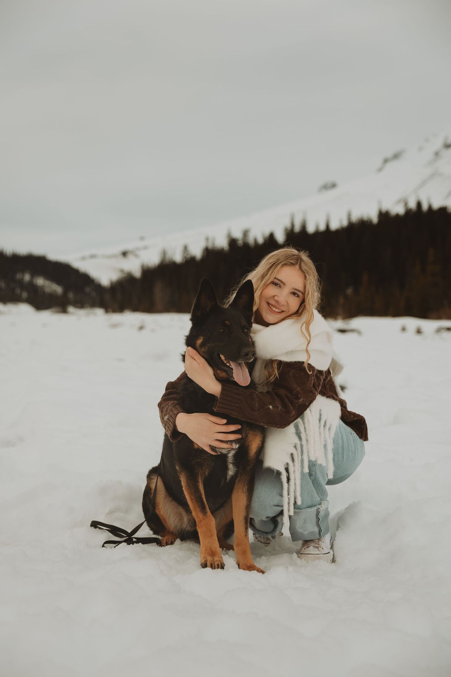 Smiling blonde woman hugs black and tan German Shepherd dog while kneeling in winter snow landscape.