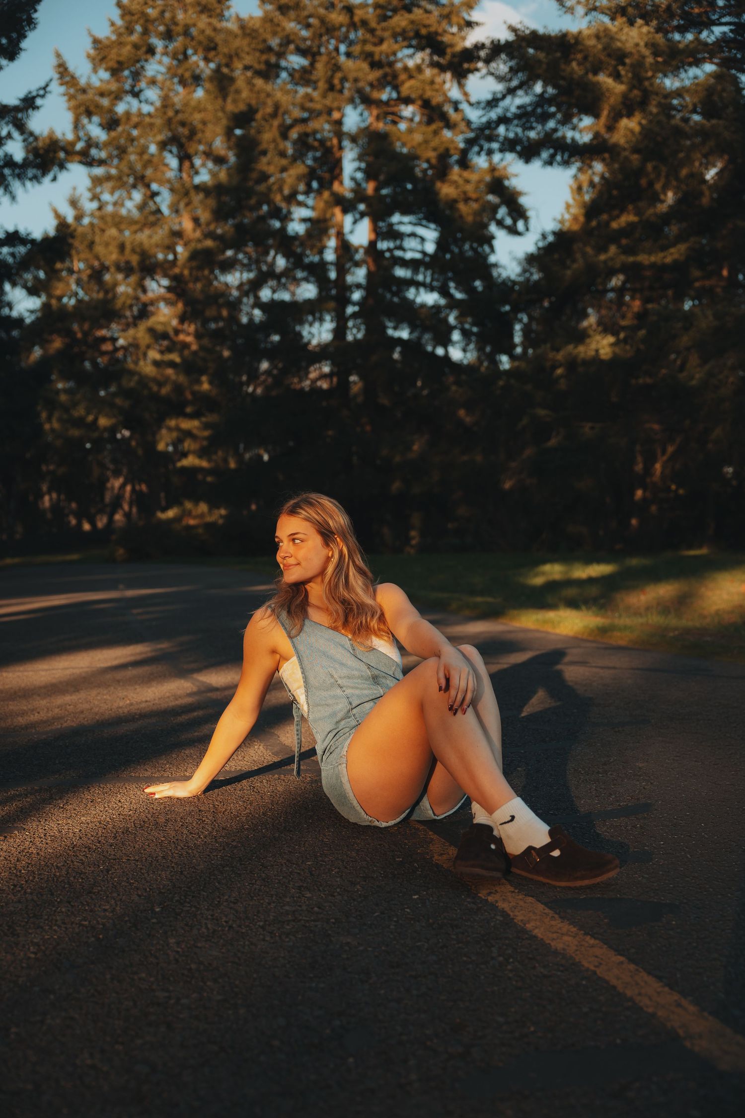 Young blonde woman sitting on a skateboard on a road at golden hour, wearing denim overalls and white socks.