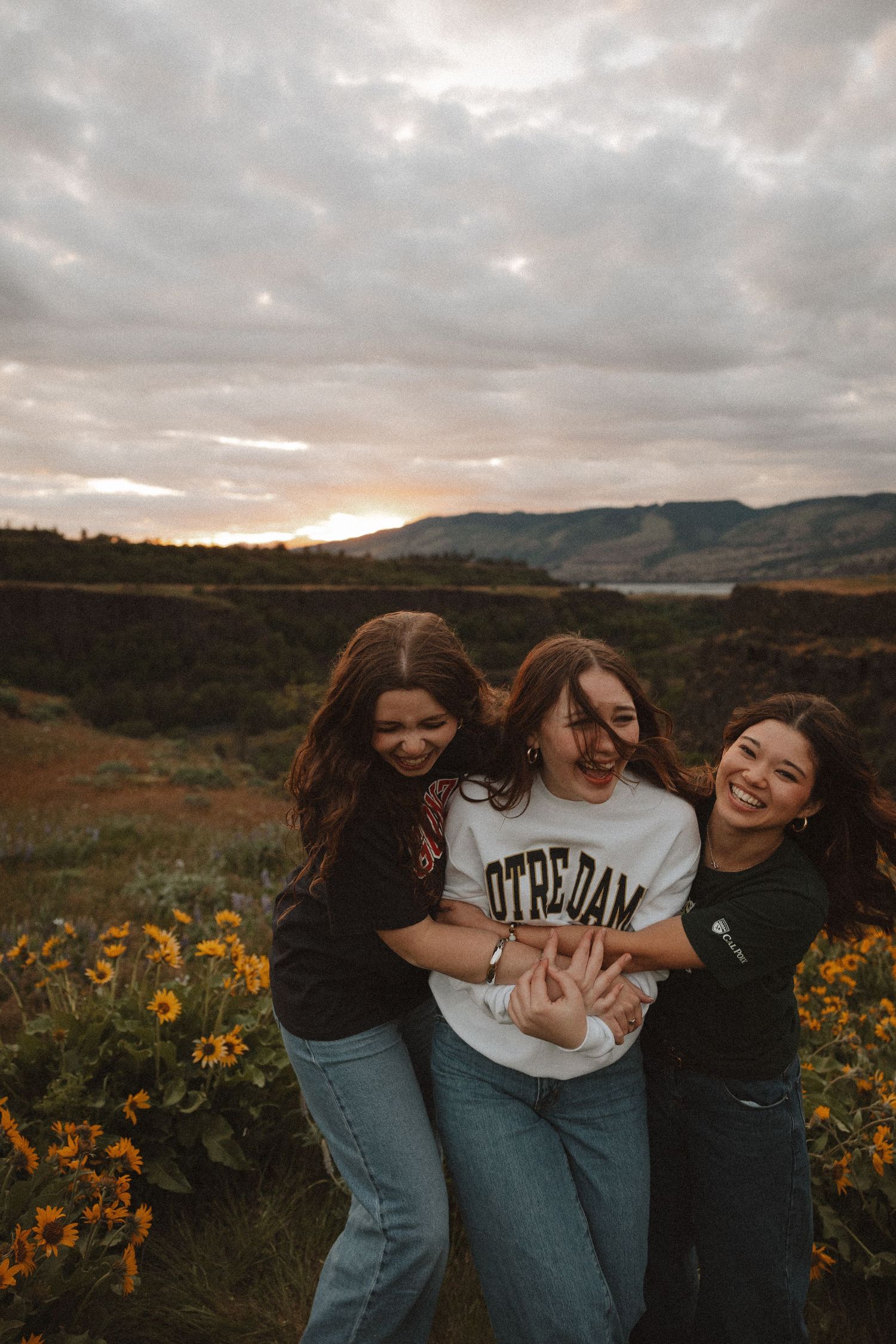 Three happy young women embracing in a yellow wildflower field at sunset with mountain views.